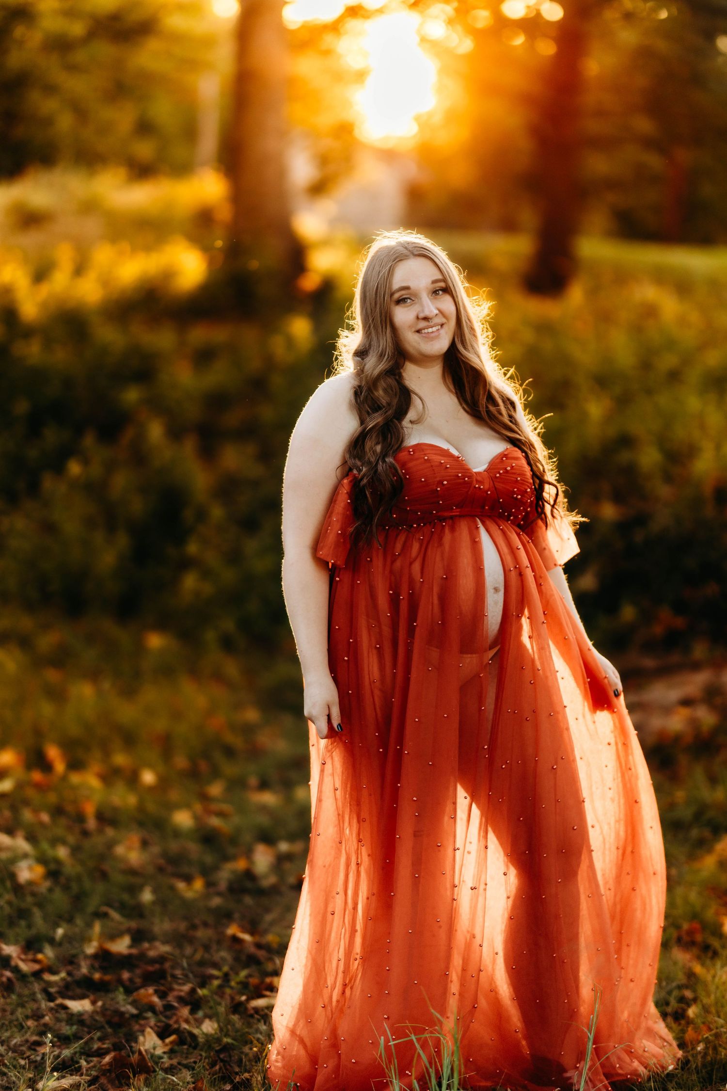 Pregnant woman in flowing rust orange tulle maternity gown posing outdoors during golden sunset.