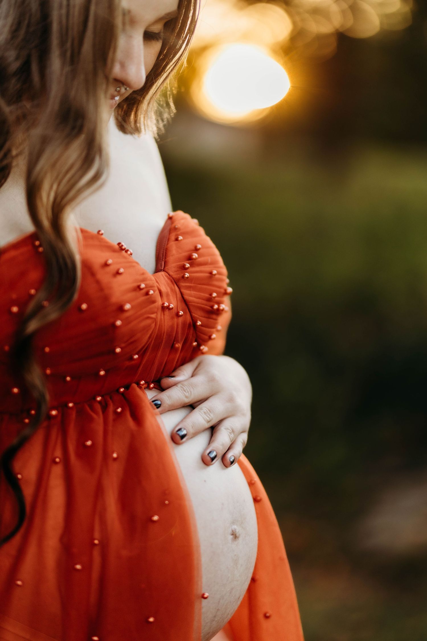 Close-up of pregnant woman in red beaded maternity dress cradling baby bump at sunset.