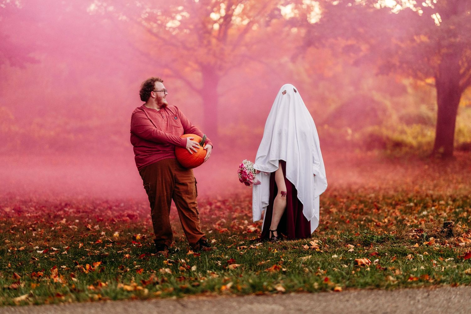 A man in a red sweater holds a pumpkin beside a person dressed as a ghost in a foggy autumn park.