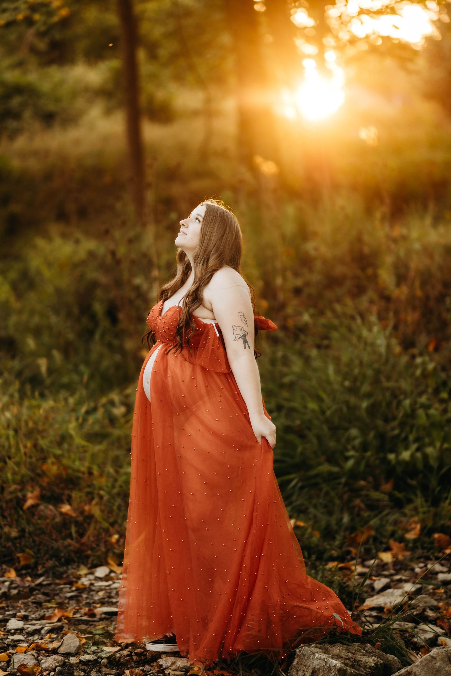 Pregnant woman in elegant rust orange tulle gown looking up at golden sunset in forest setting.