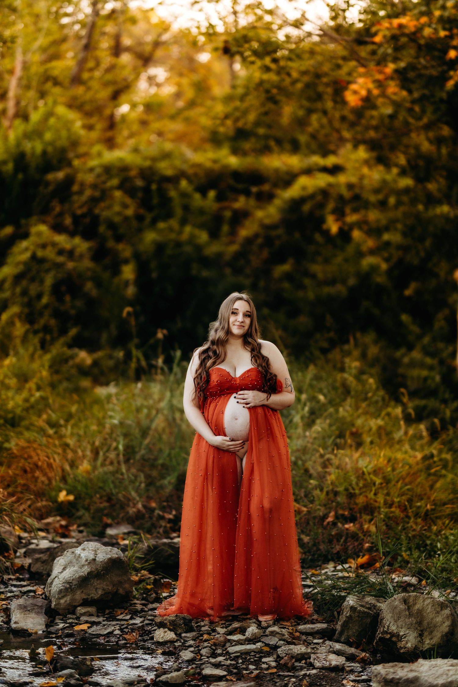 Pregnant woman in flowing red tulle maternity dress standing on rocks near stream in autumn forest.