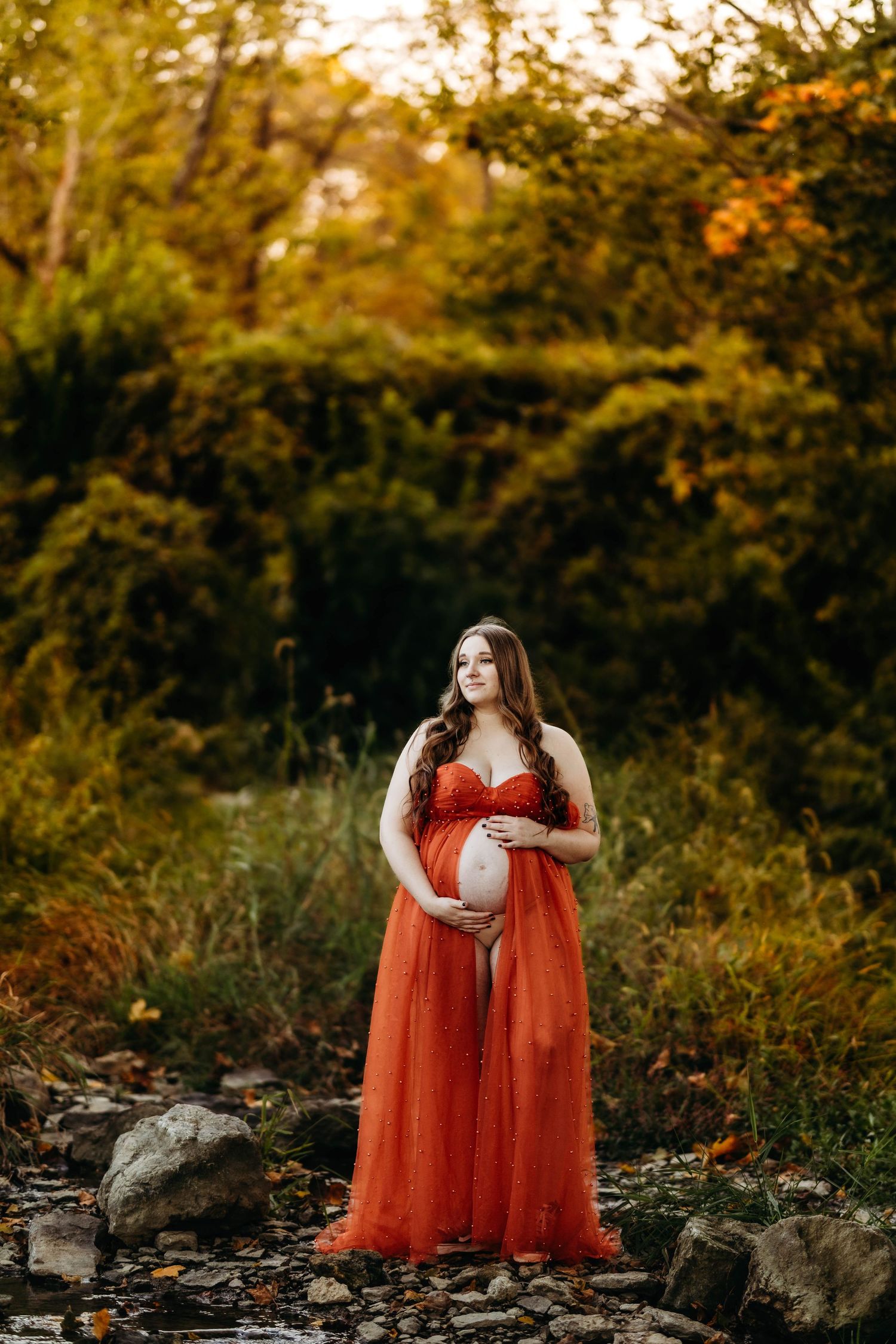 Pregnant woman in open red tulle maternity gown cradling bare baby bump in autumn nature setting.