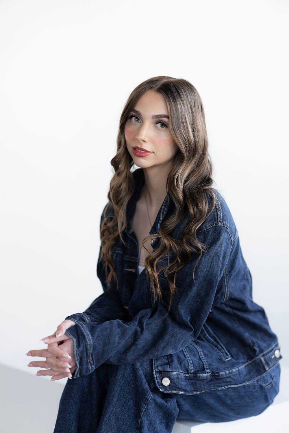 Young woman with long wavy hair wearing oversized denim jacket sitting against white background.