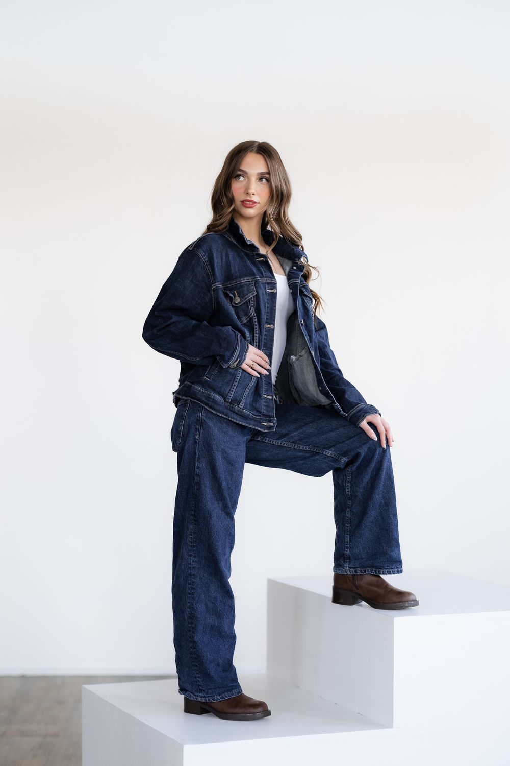 Young woman in dark denim jacket and wide-leg jeans posing on white steps with brown boots.