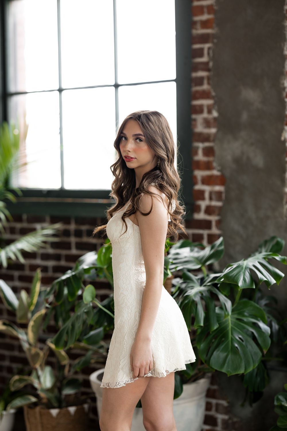 Young woman in white lace mini dress posing near window with tropical plants and exposed brick wall.