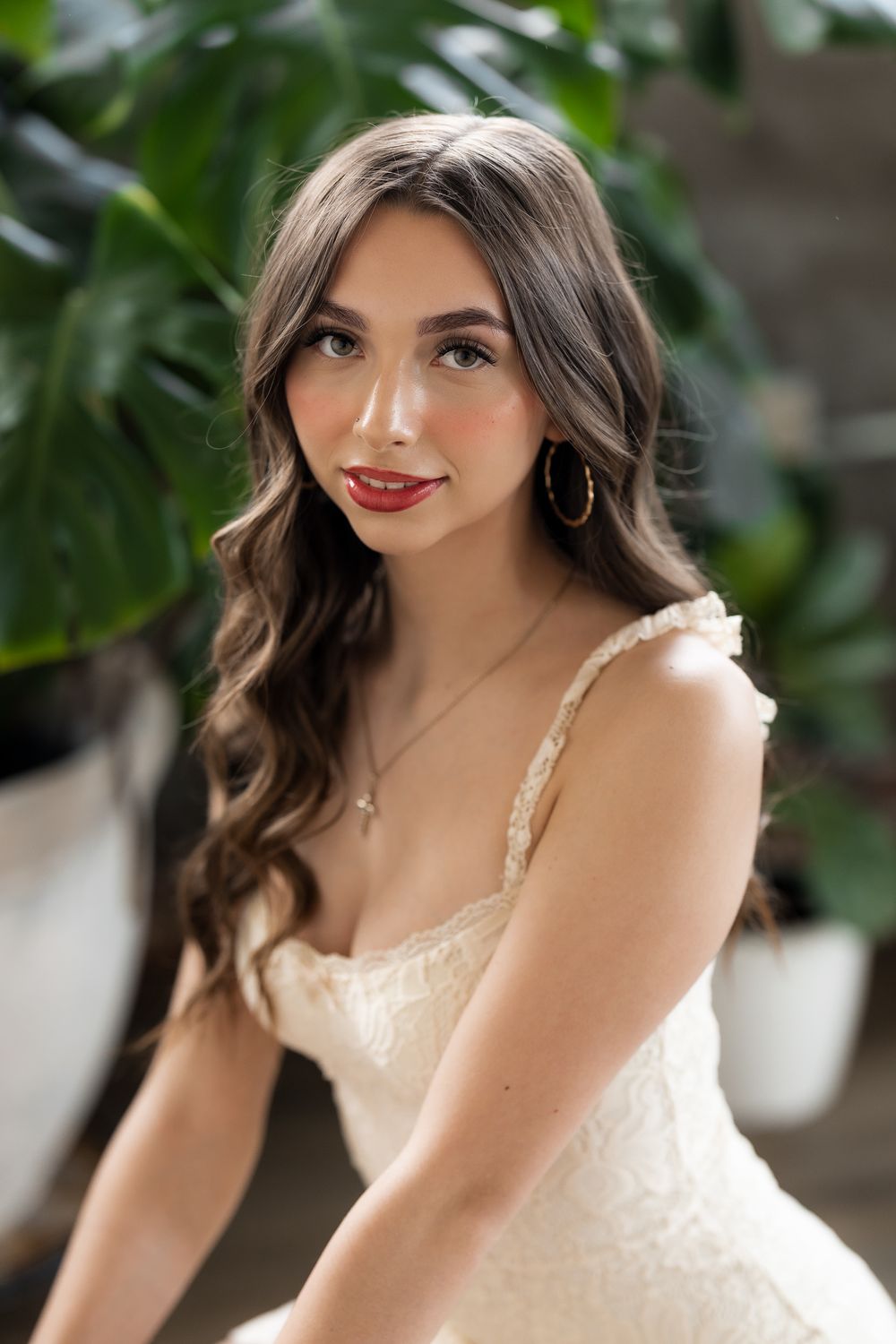 Young woman with long wavy dark hair in white lace dress, gold earrings, and red lips poses near tropical plants.