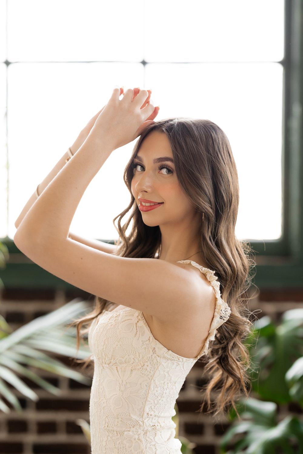 A young woman in a white lace dress poses gracefully with her hand raised, long wavy hair flowing, smiling softly near a bright window.