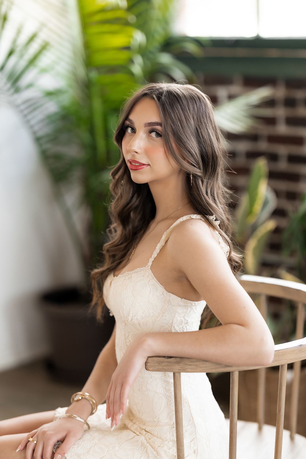 Young woman in white lace dress sitting gracefully on wooden chair with tropical plants in background.