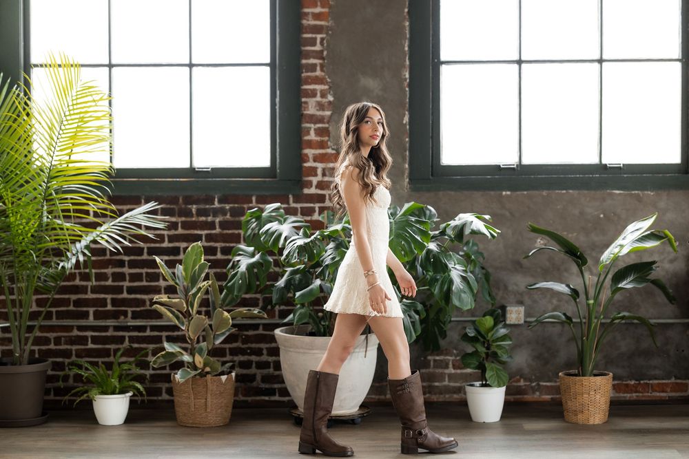 Woman in white dress and brown boots walks among lush indoor plants against a brick wall backdrop.