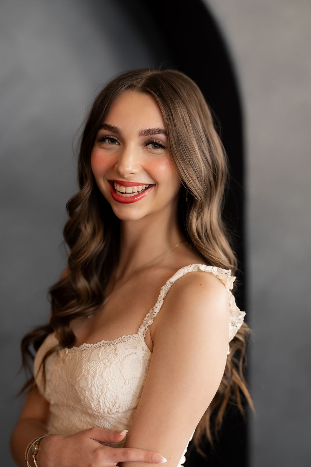 Smiling young woman with long wavy brown hair wearing a white lace dress poses against a gray backdrop.