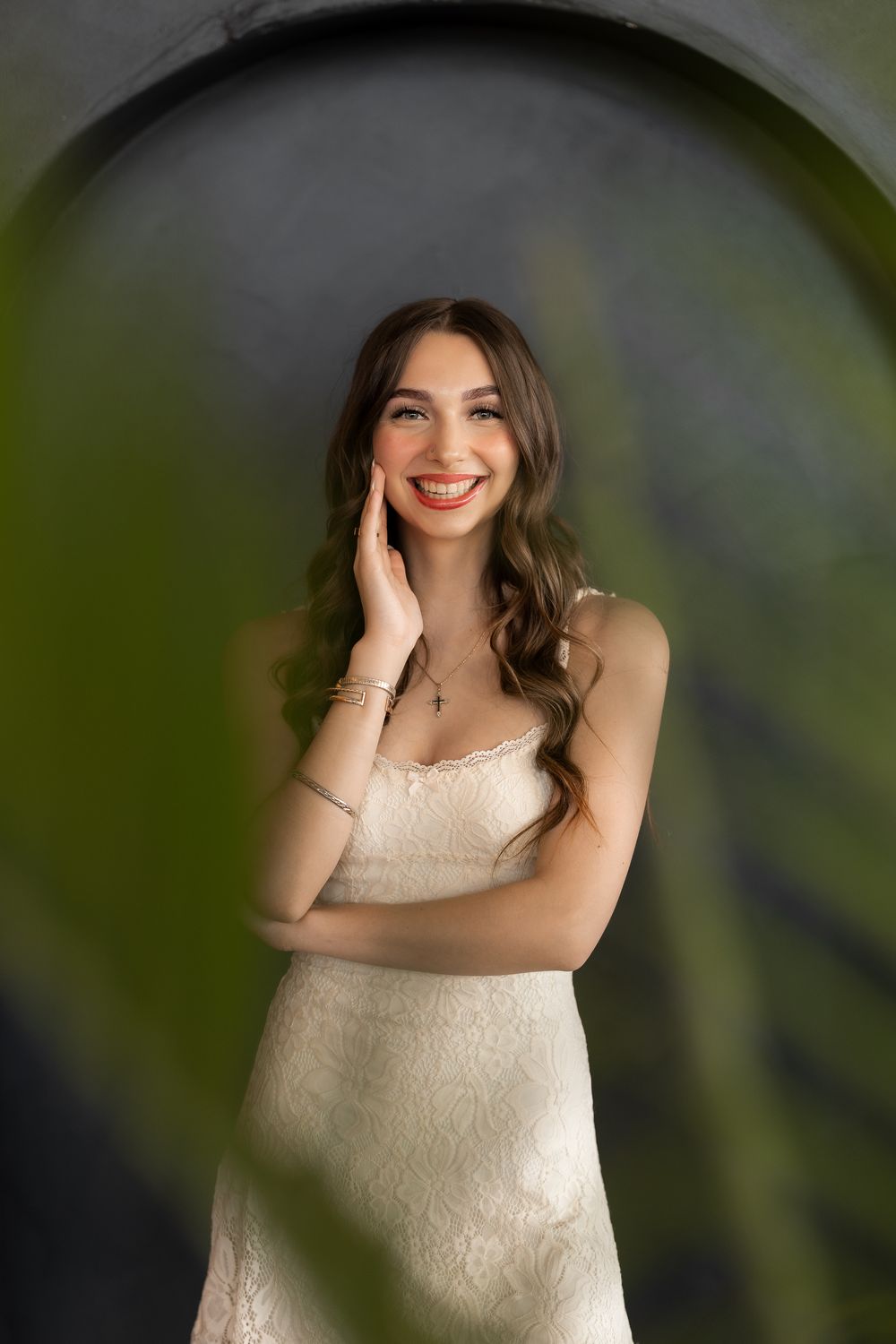 Smiling young woman in white lace dress posing gracefully with hand on cheek against dark arched backdrop.