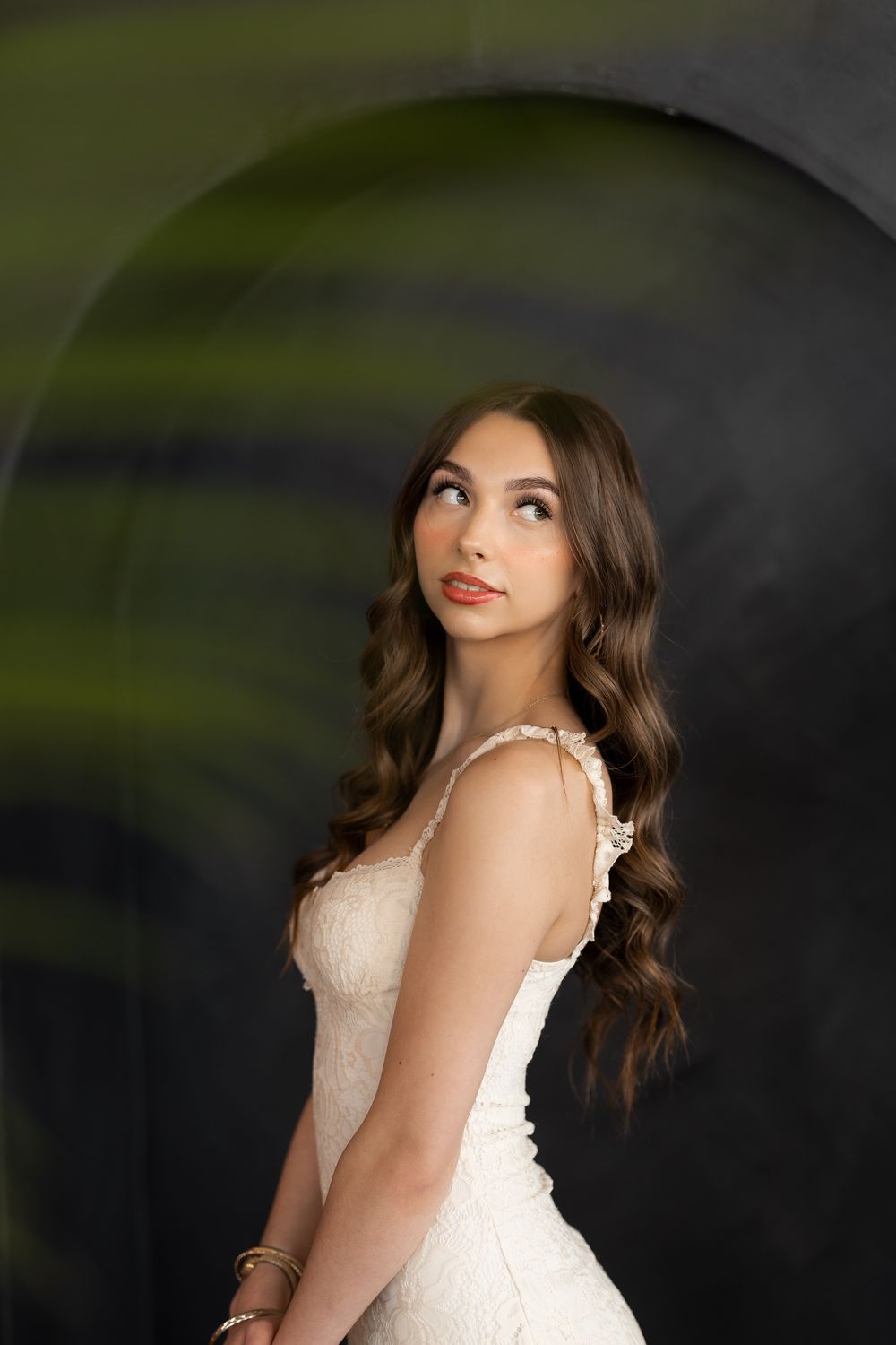 Young woman in white lace dress with long wavy hair gazes upward, posed against dark arch backdrop.