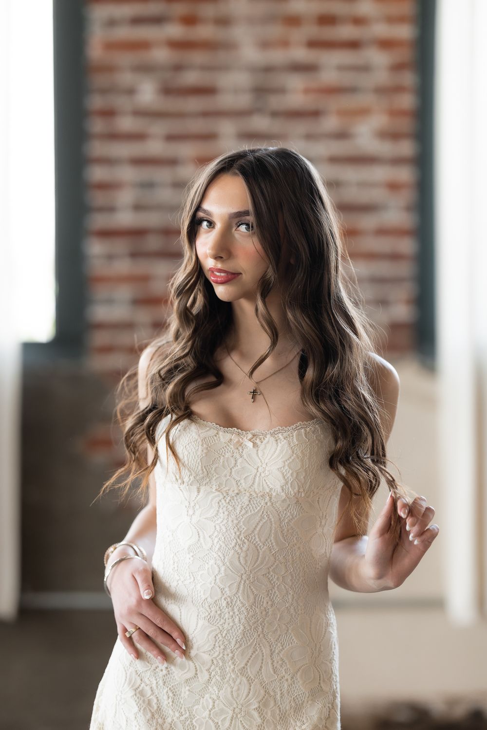 Elegant young woman in strapless white lace dress posing against exposed brick wall interior.