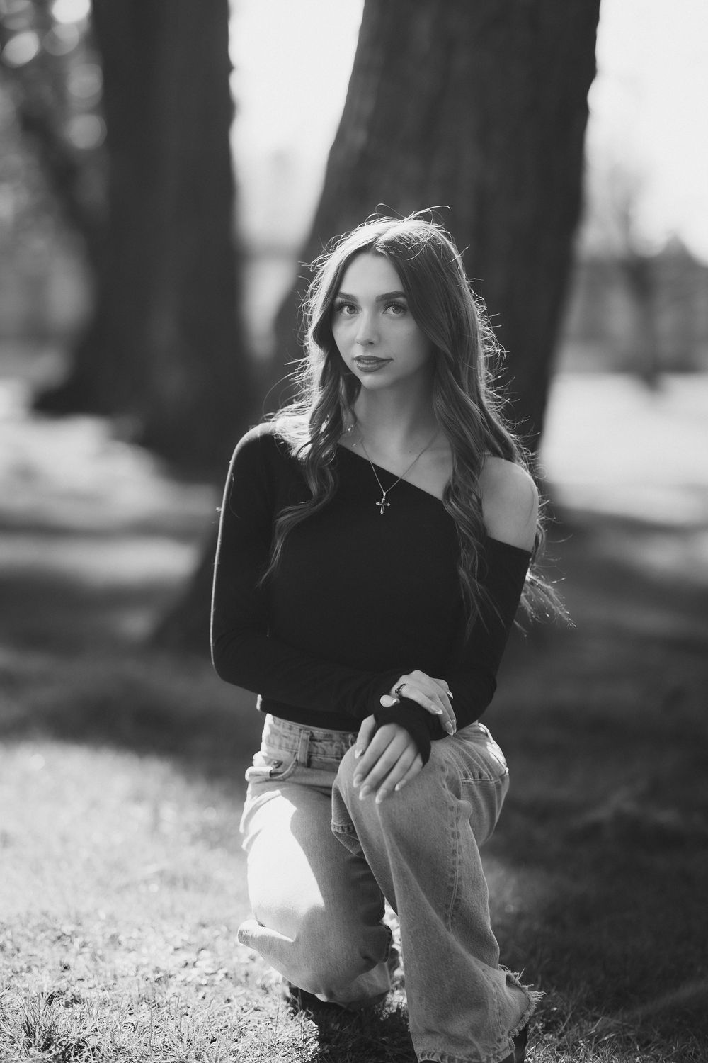 Young woman with long wavy hair poses outdoors in black off-shoulder top and jeans, black and white portrait.