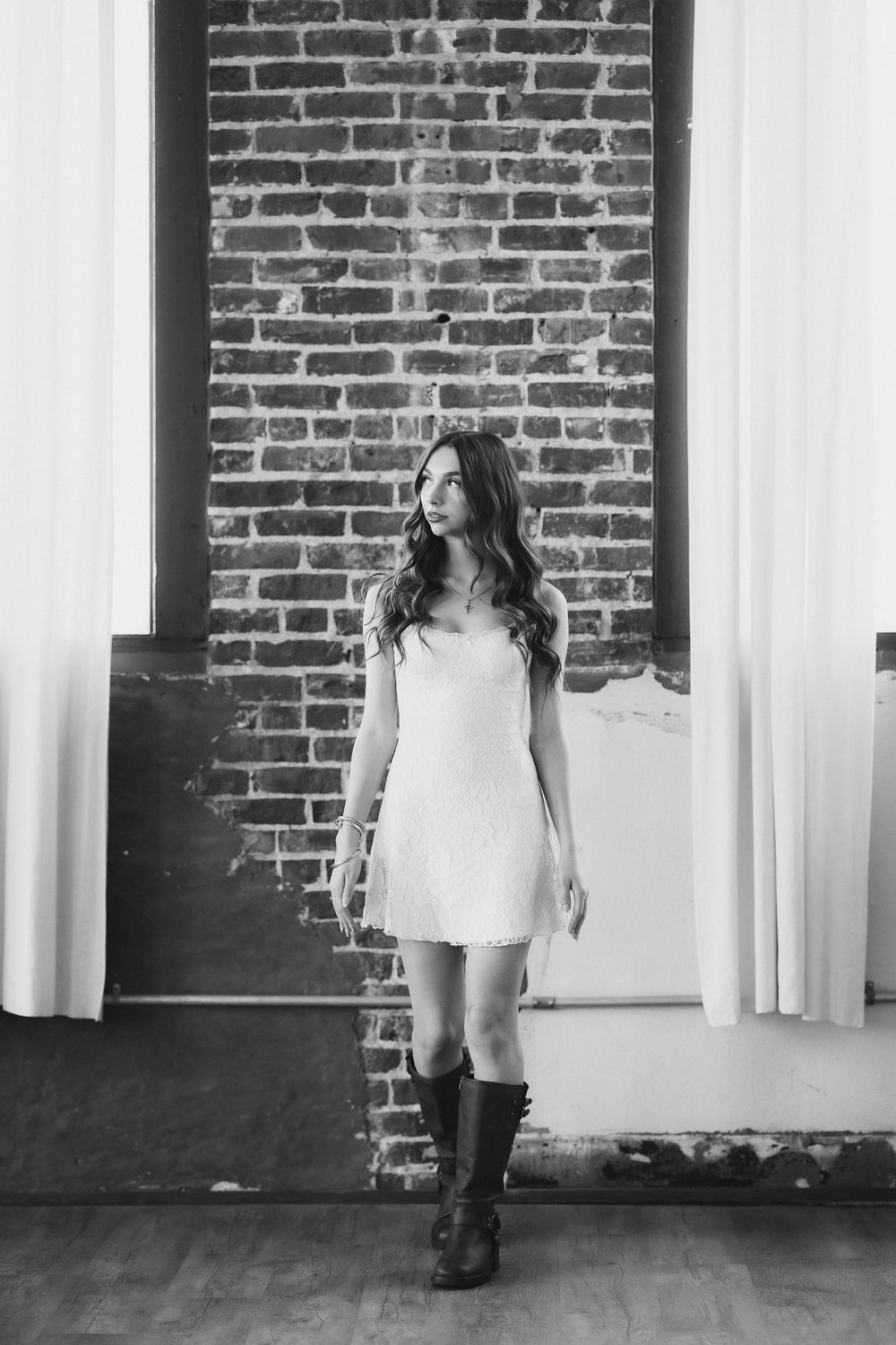 Black and white photo of young woman in white mini dress and boots by brick wall.