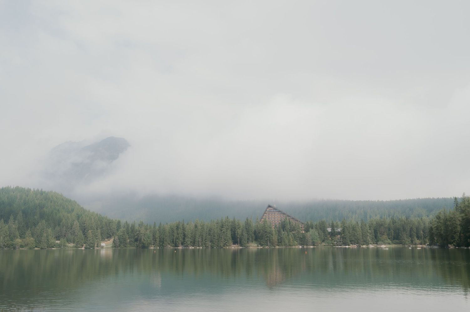 Misty lake reflecting dense pine forest with a rustic cabin and fog-covered mountains in the background.