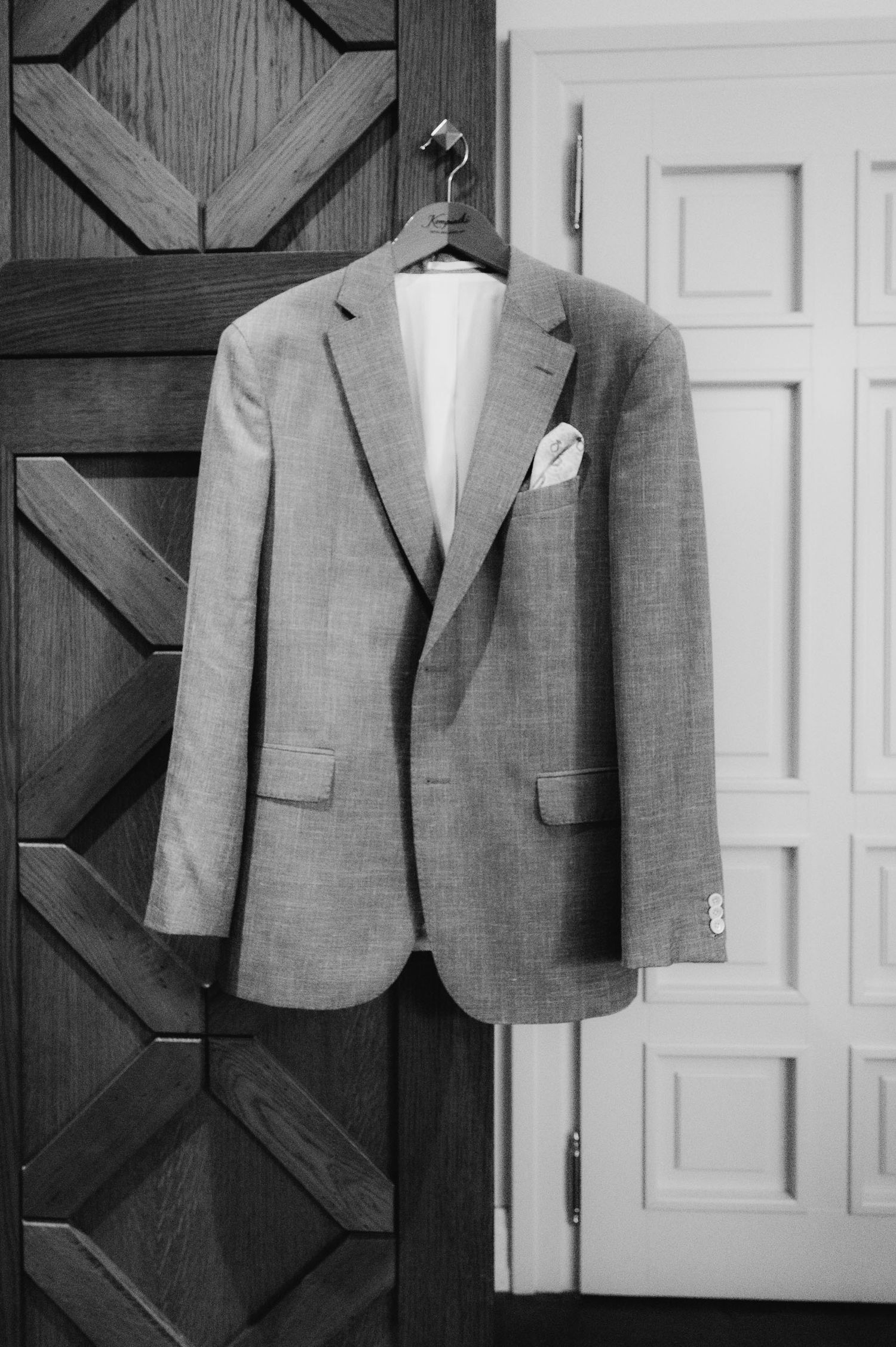 A classic men's blazer with pocket square hangs on a wooden door in black and white photography.