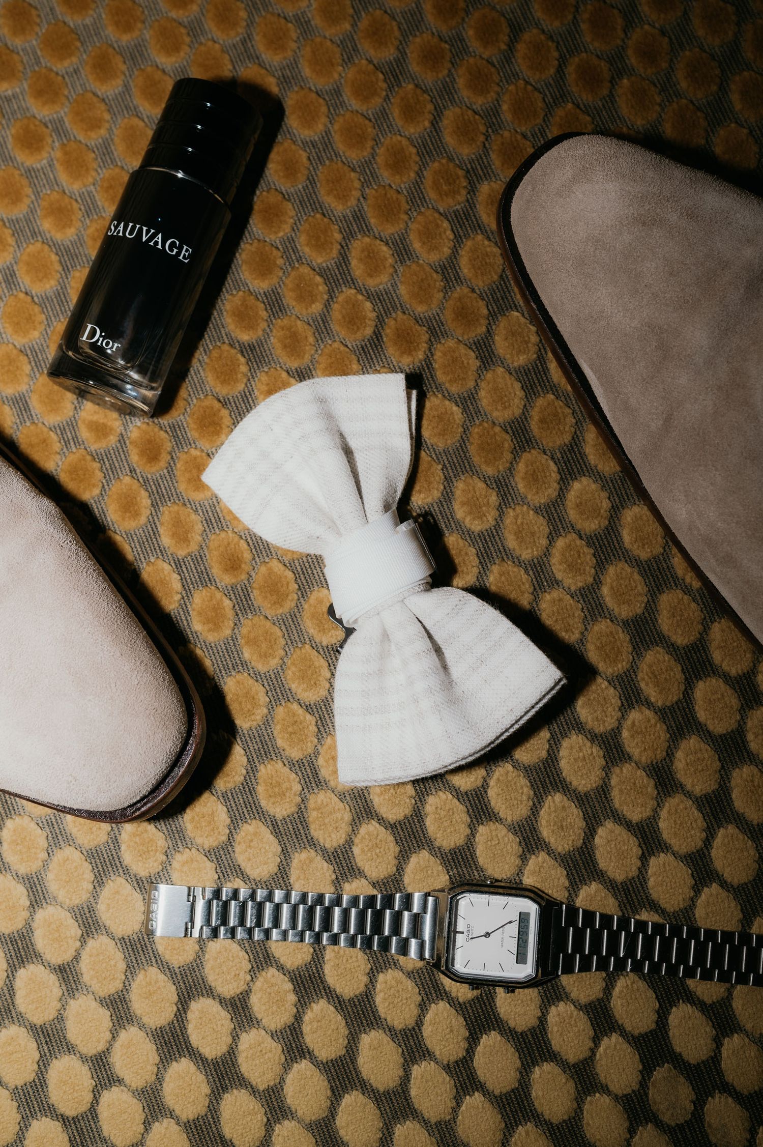 Flat lay of Dior Sauvage cologne, white bow tie, silver watch, and suede shoes on patterned carpet.