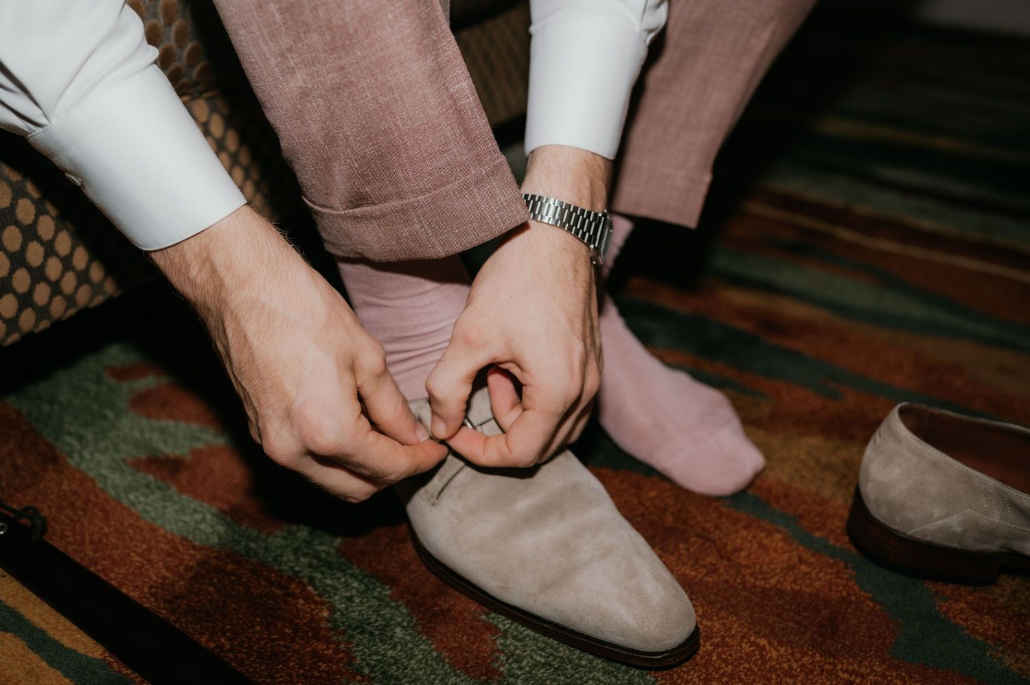 Man in pink trousers and silver watch tying suede dress shoes on a colorful patterned carpet.