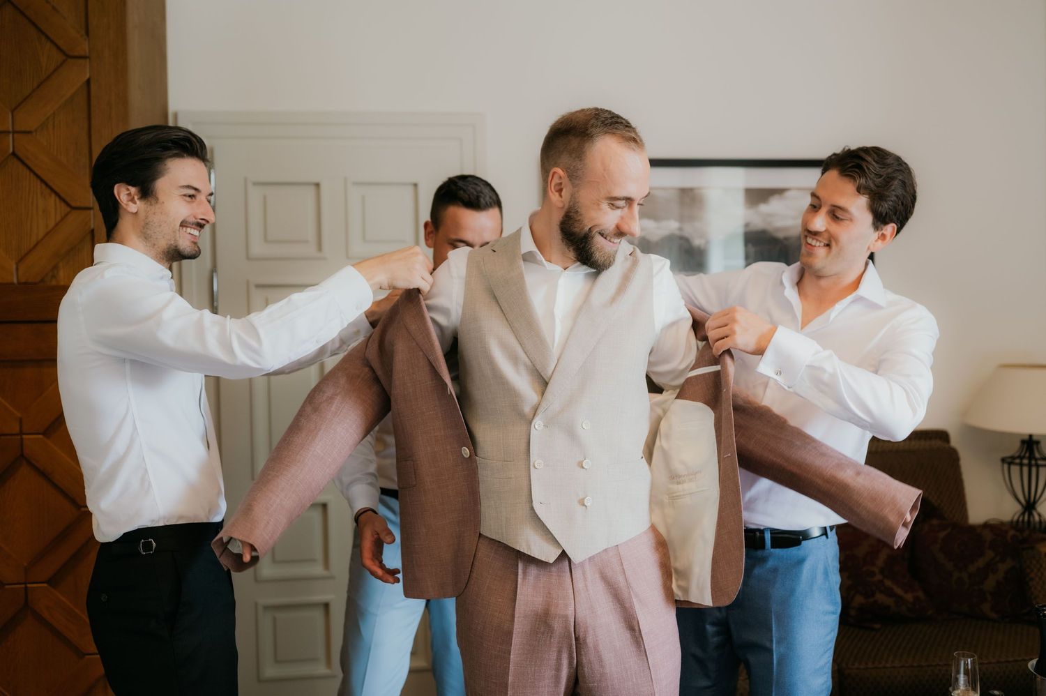 Groomsmen helping groom put on brown suit jacket while smiling during wedding preparation indoors.