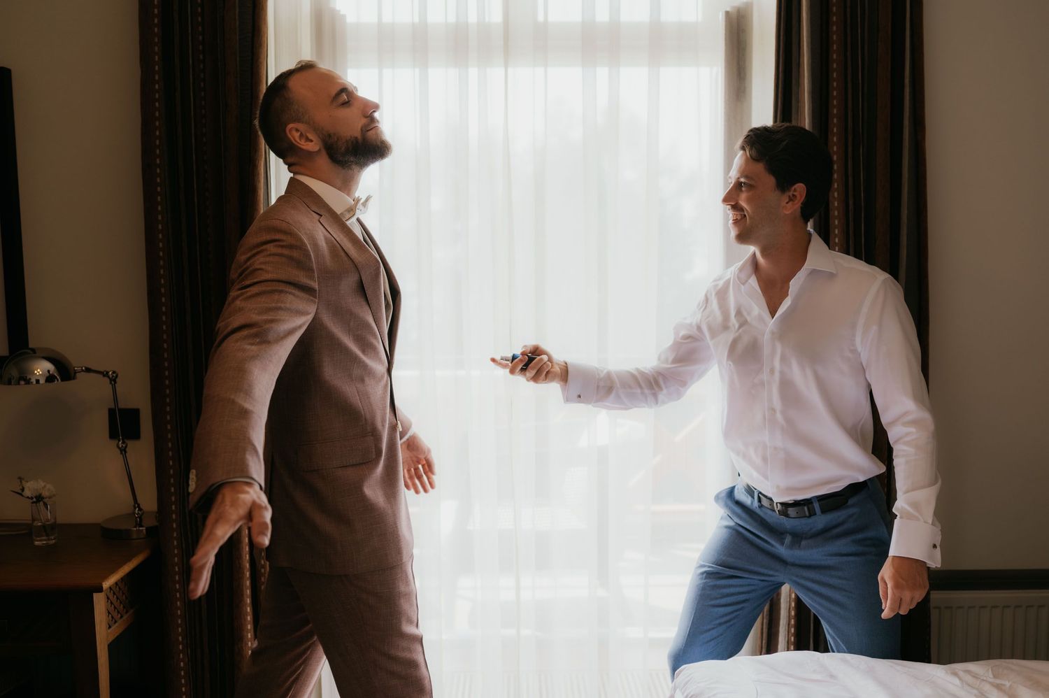 Two men in formal attire laugh and joke around in a hotel room, one in a brown suit and one in a white shirt.