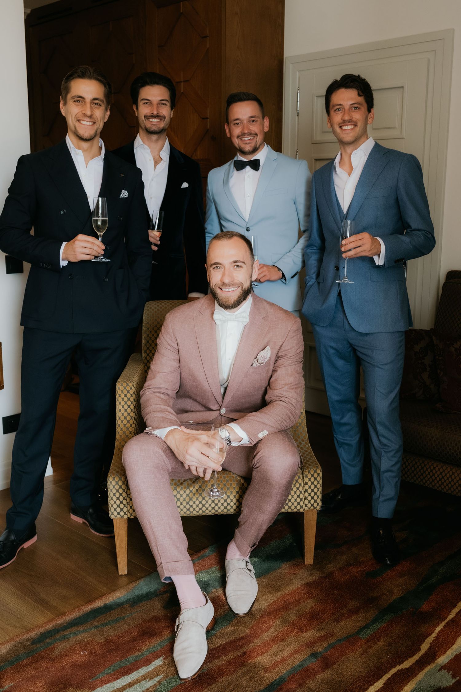 Five groomsmen in suits pose together, with groom seated in pink suit holding champagne glasses at wedding celebration.