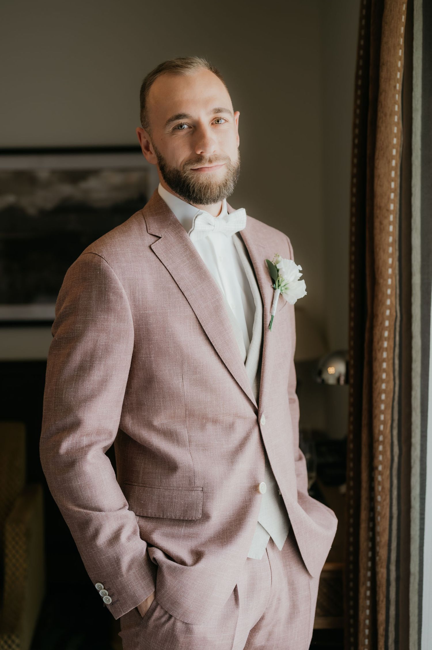 A dapper groom in a dusty rose suit with white boutonniere and bow tie poses confidently with hands in pockets.