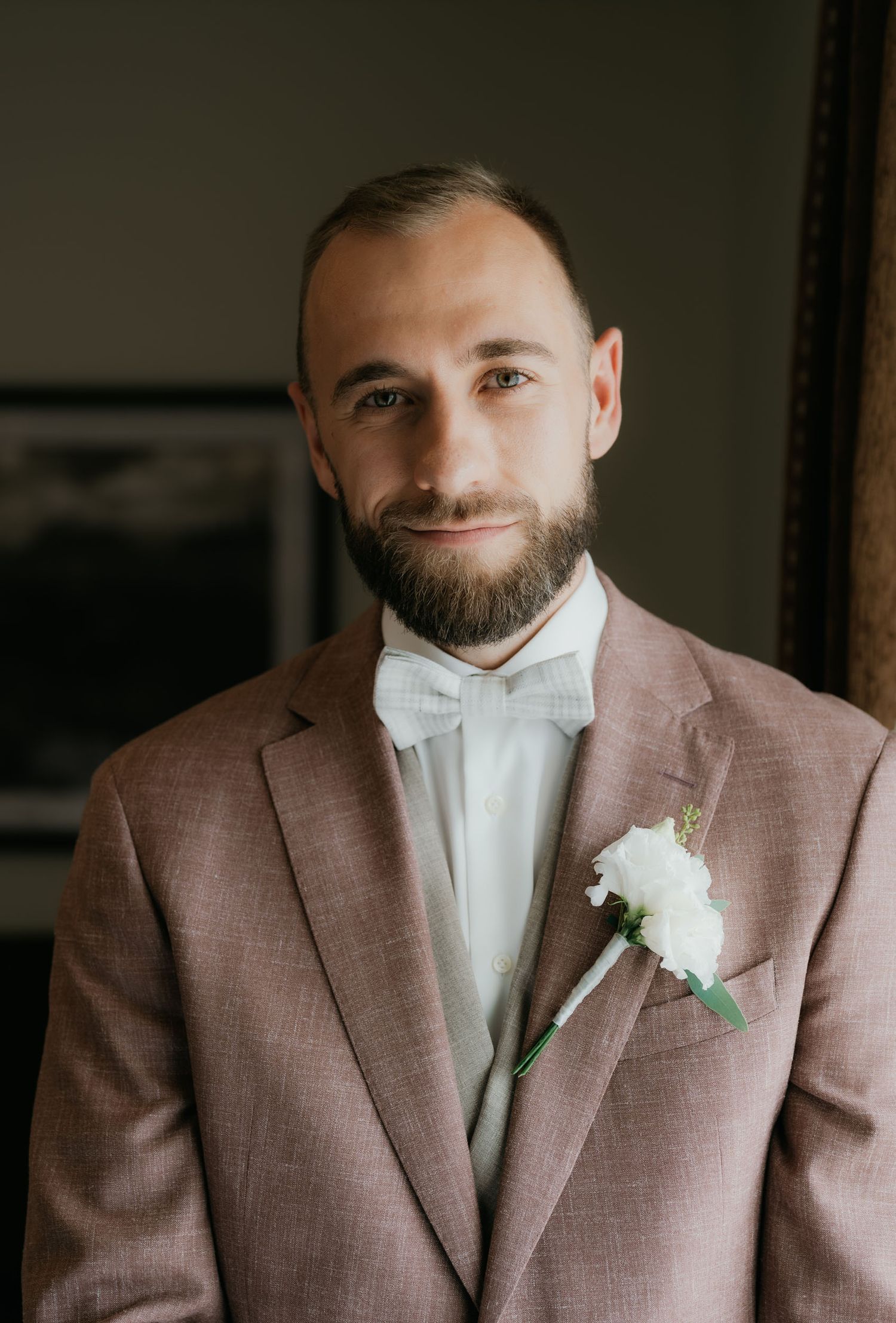 Groom in pink suit with white boutonniere and bow tie smiling for formal wedding portrait photo.