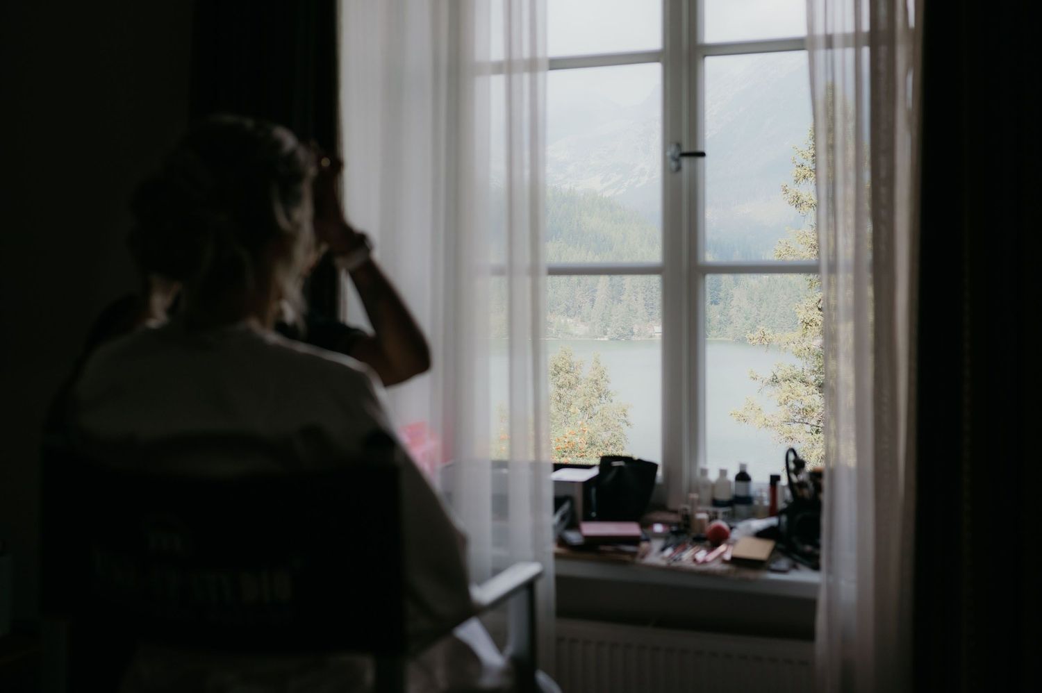 Person sitting by a bright window with sheer curtains, looking outside at a scenic view with trees and water.