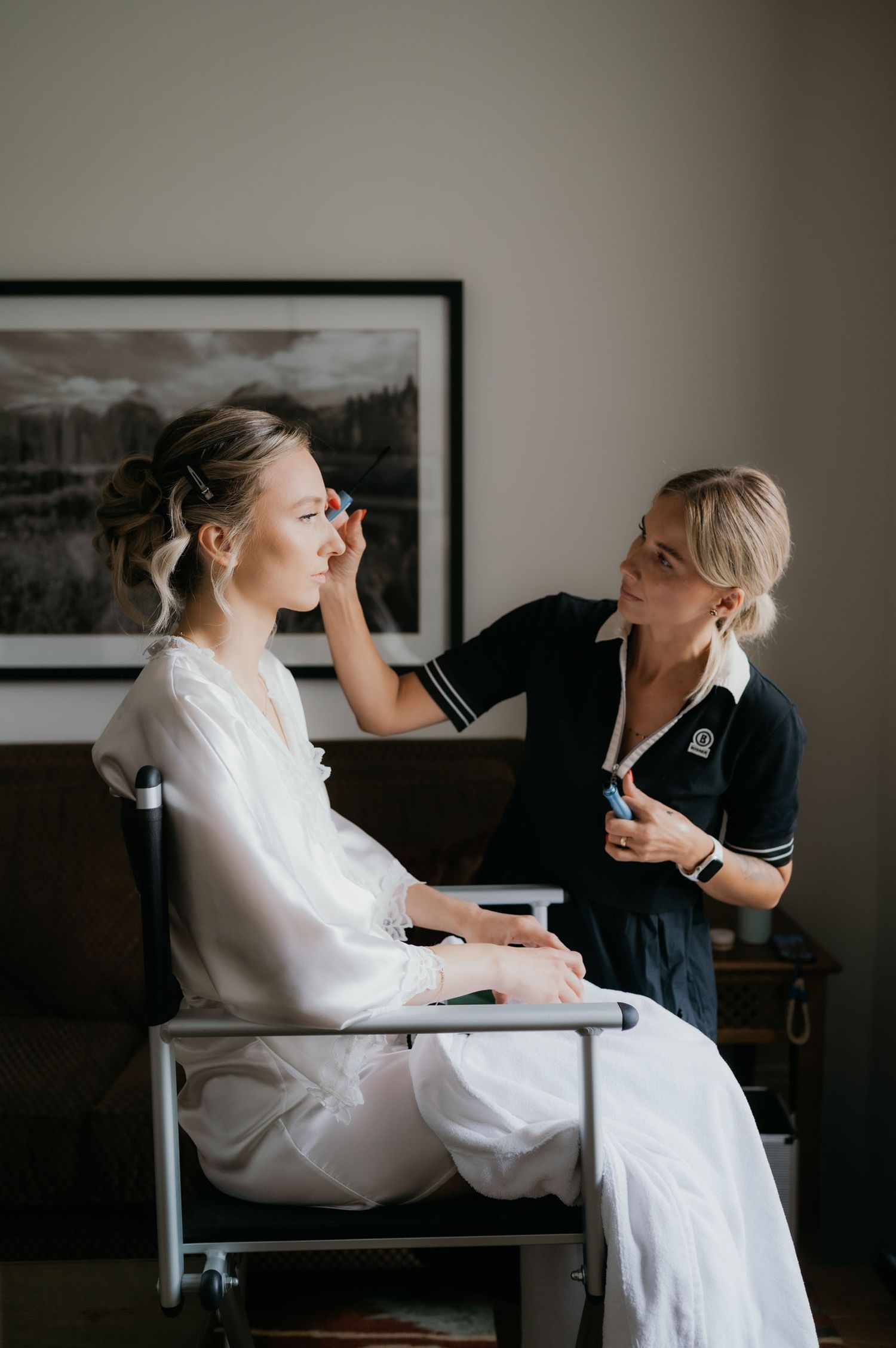 A makeup artist applies eye makeup to a bride in a white robe seated in a chair before her wedding.