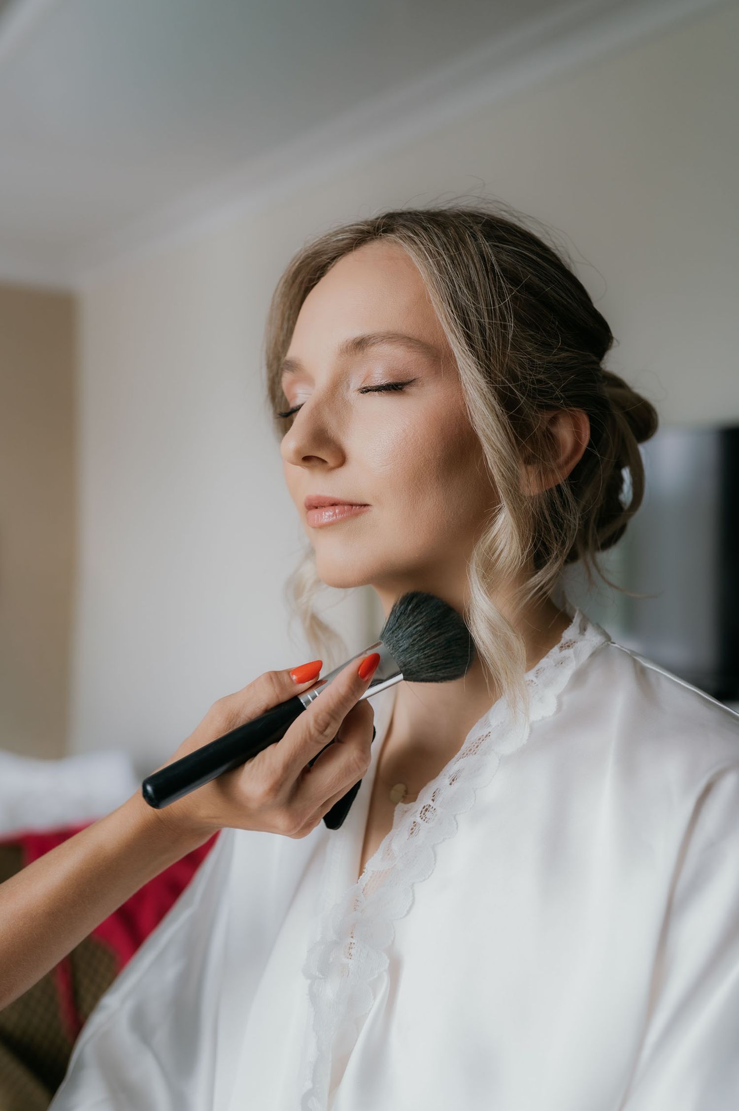 A makeup artist applies powder blush to a bride in a white robe with her eyes closed before her wedding.