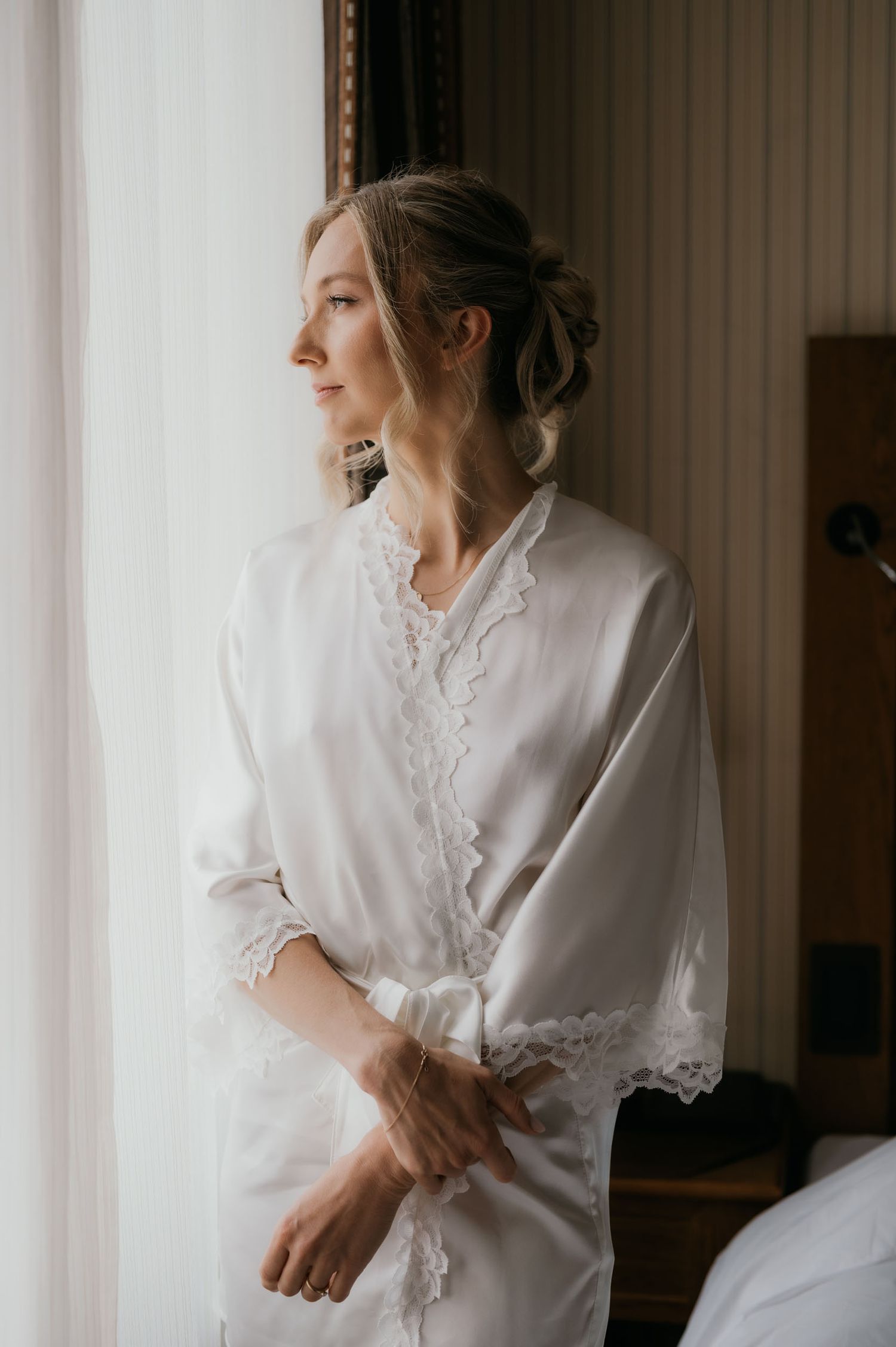 Bride in white lace robe gazing out window, elegant updo hairstyle, soft natural light, bridal getting ready photo.