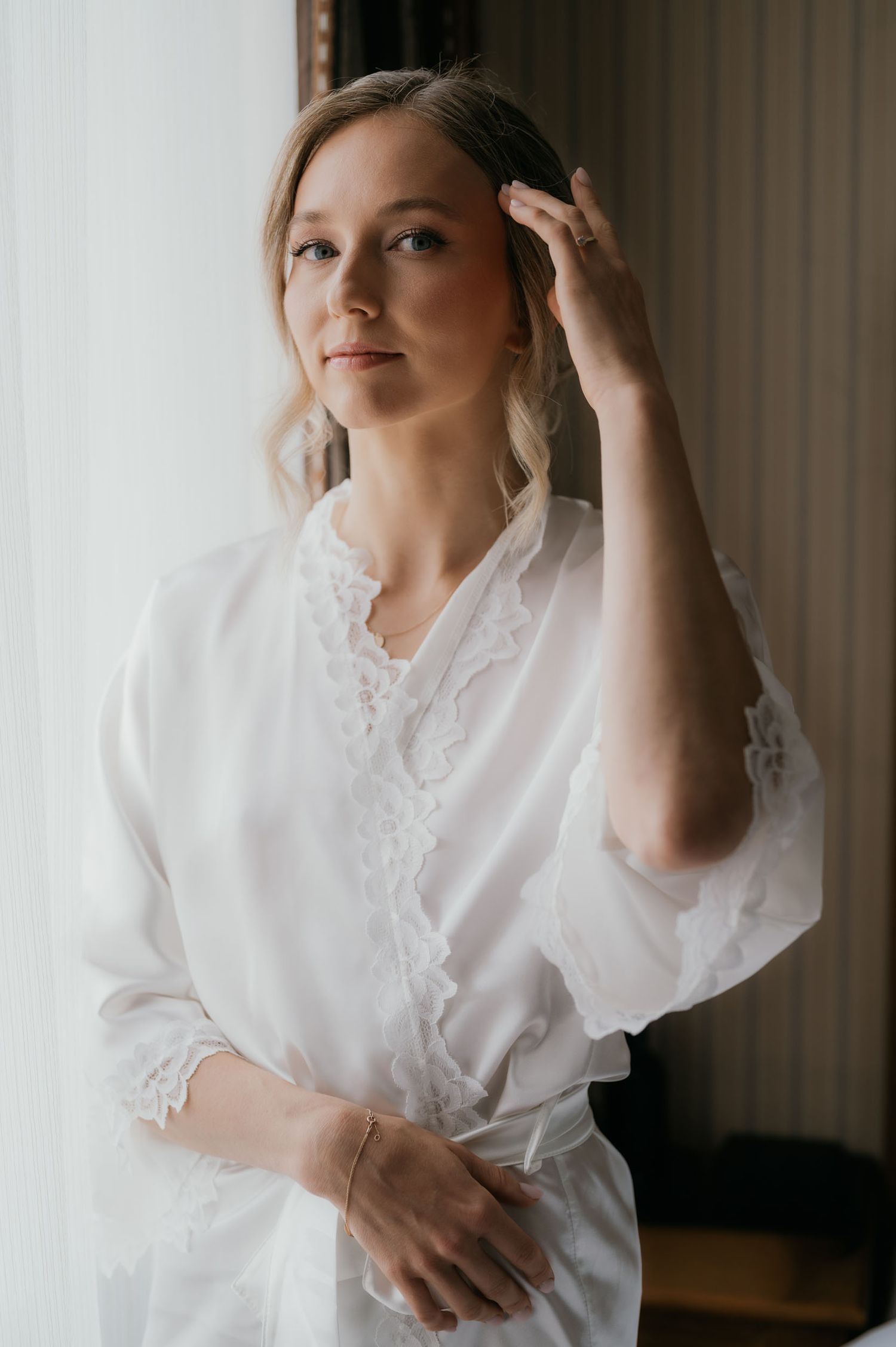 A blonde woman in a white lace robe poses gracefully near a bright window, touching her hair softly.