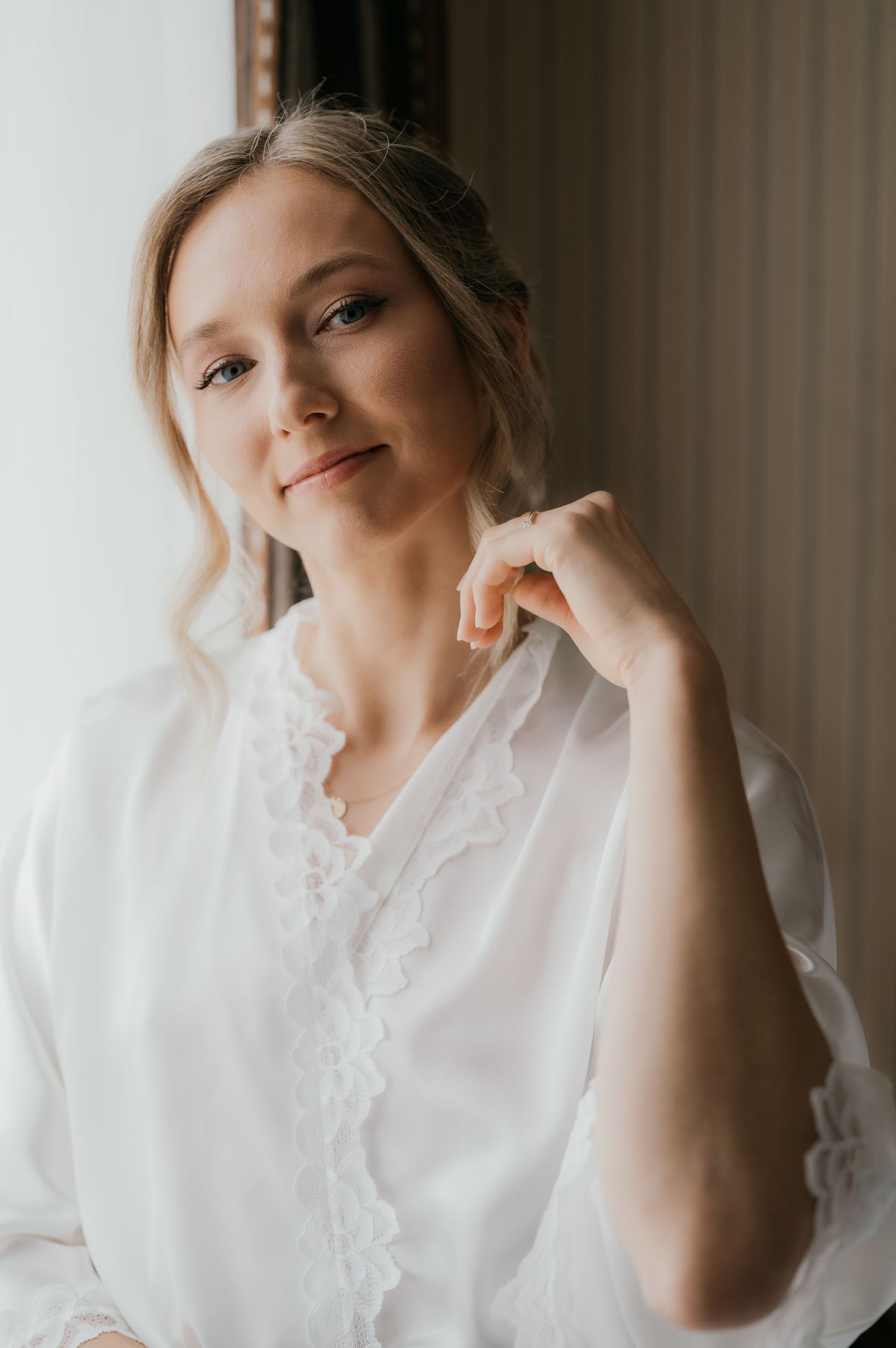 A blonde woman in a white lace robe poses gracefully near a window with soft natural light.