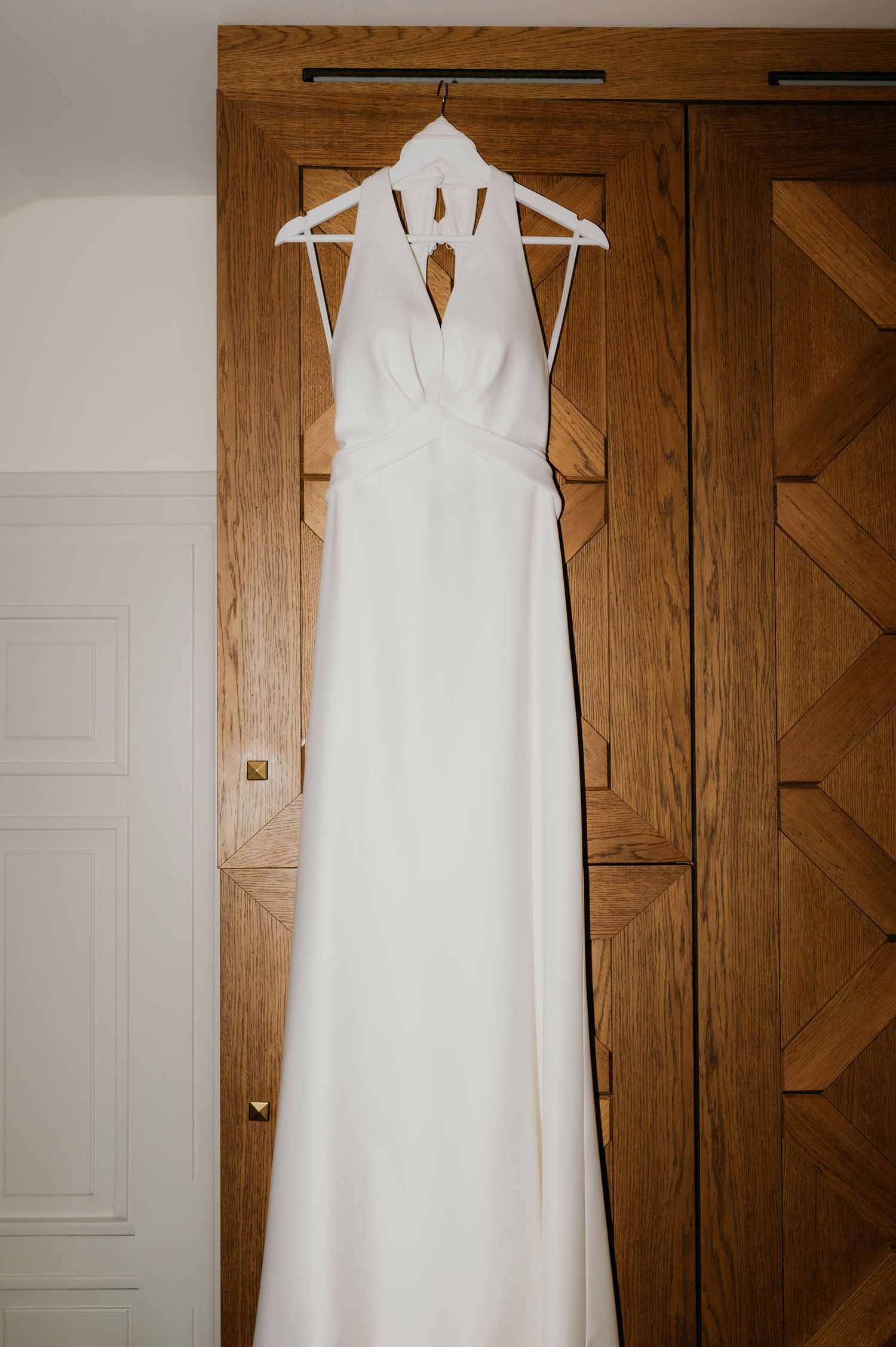 Elegant white halter neck wedding dress hanging on a wooden wardrobe door on a white hanger.