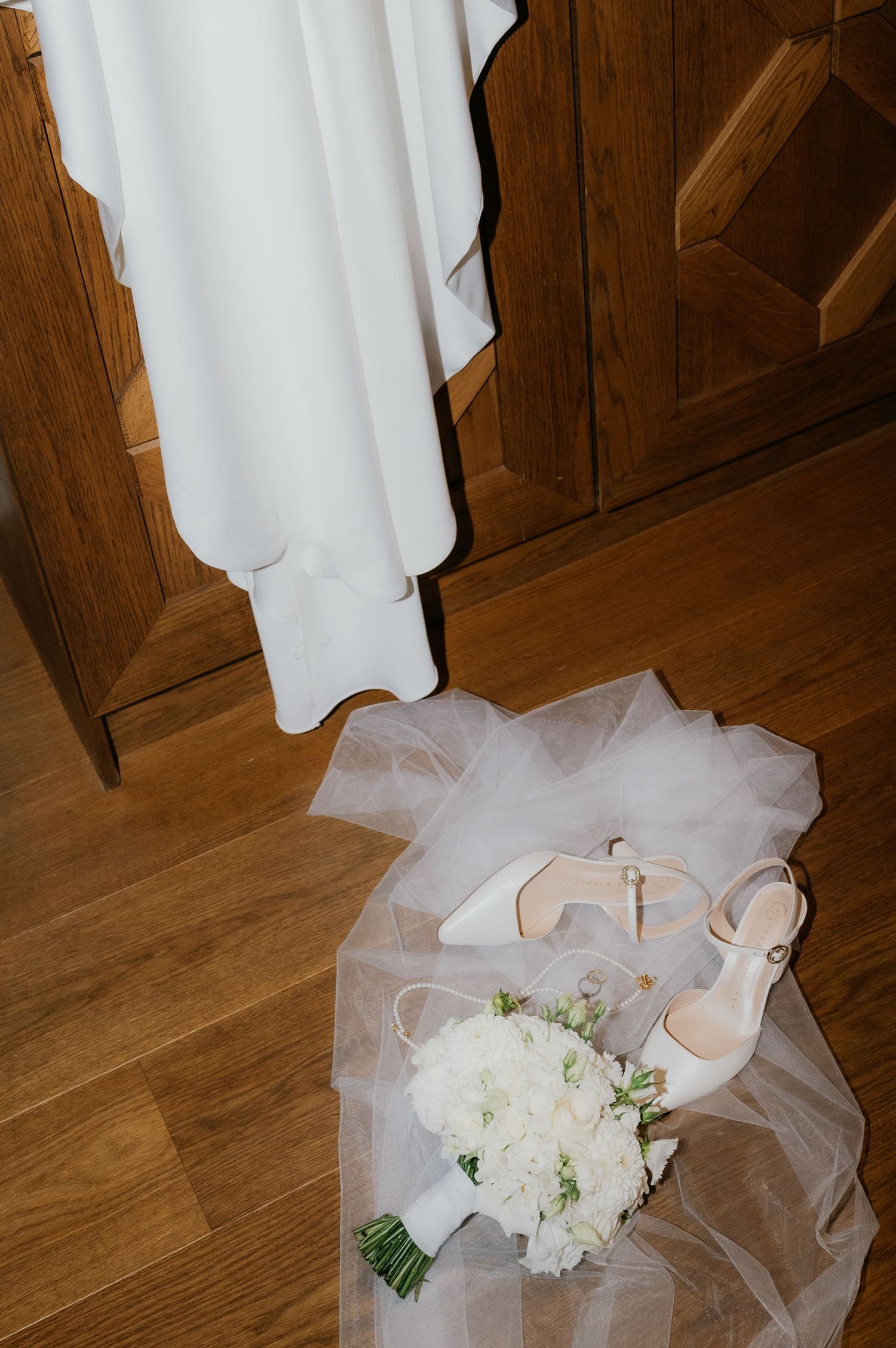Bridal flat lay with white heels, tulle veil, and white floral bouquet on hardwood floor near wedding dress.