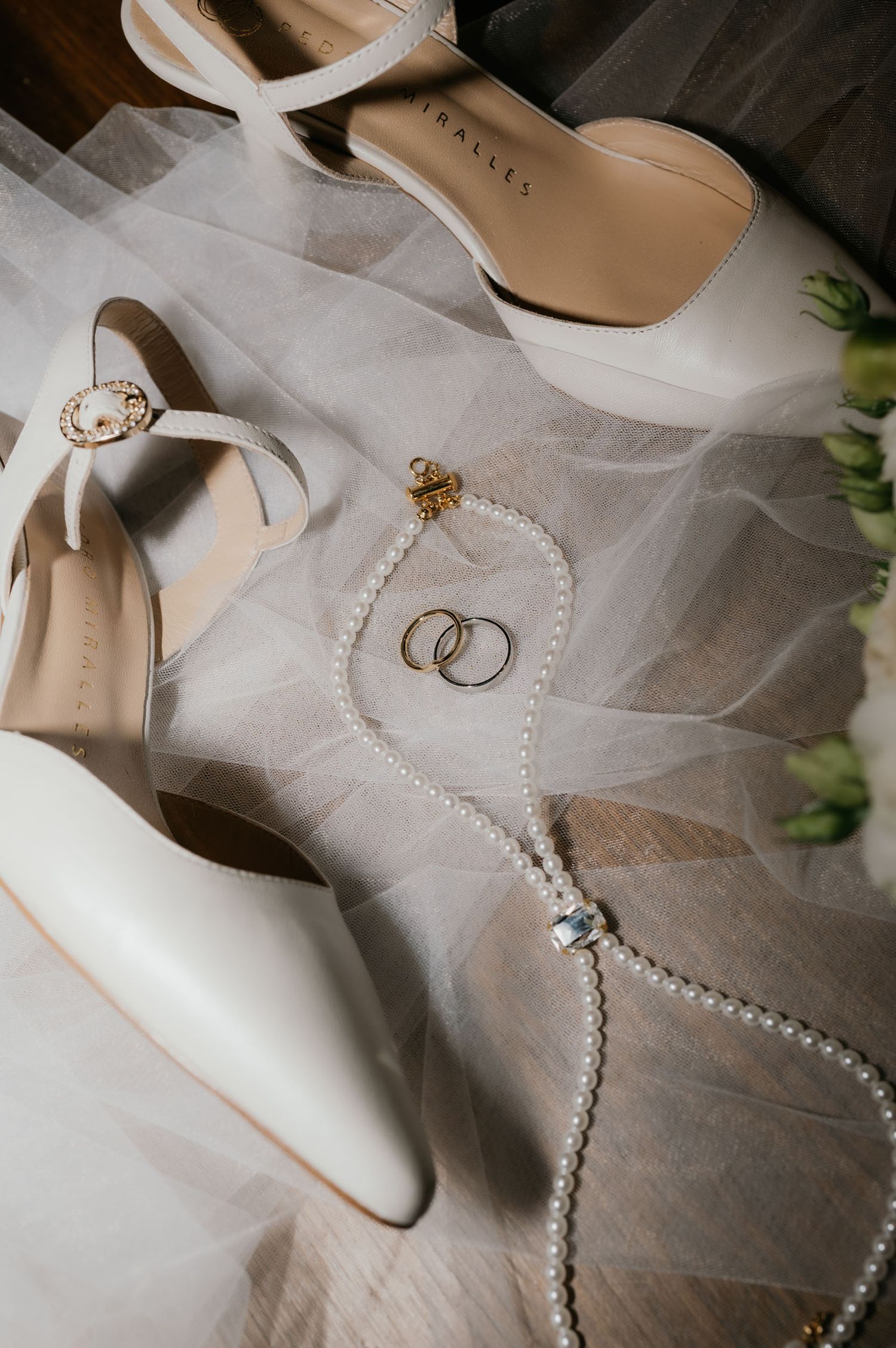 Elegant white bridal heels with pearl necklace and gold wedding rings on a soft tulle veil backdrop.