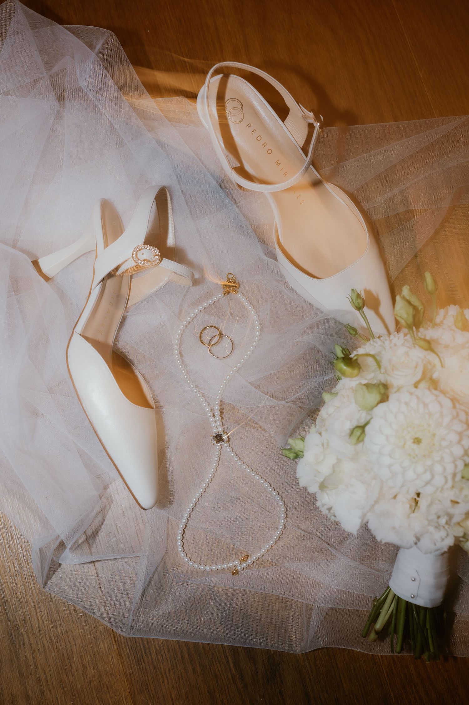Elegant bridal flat lay with white heels, pearl necklace, gold rings, and white floral bouquet on sheer veil.