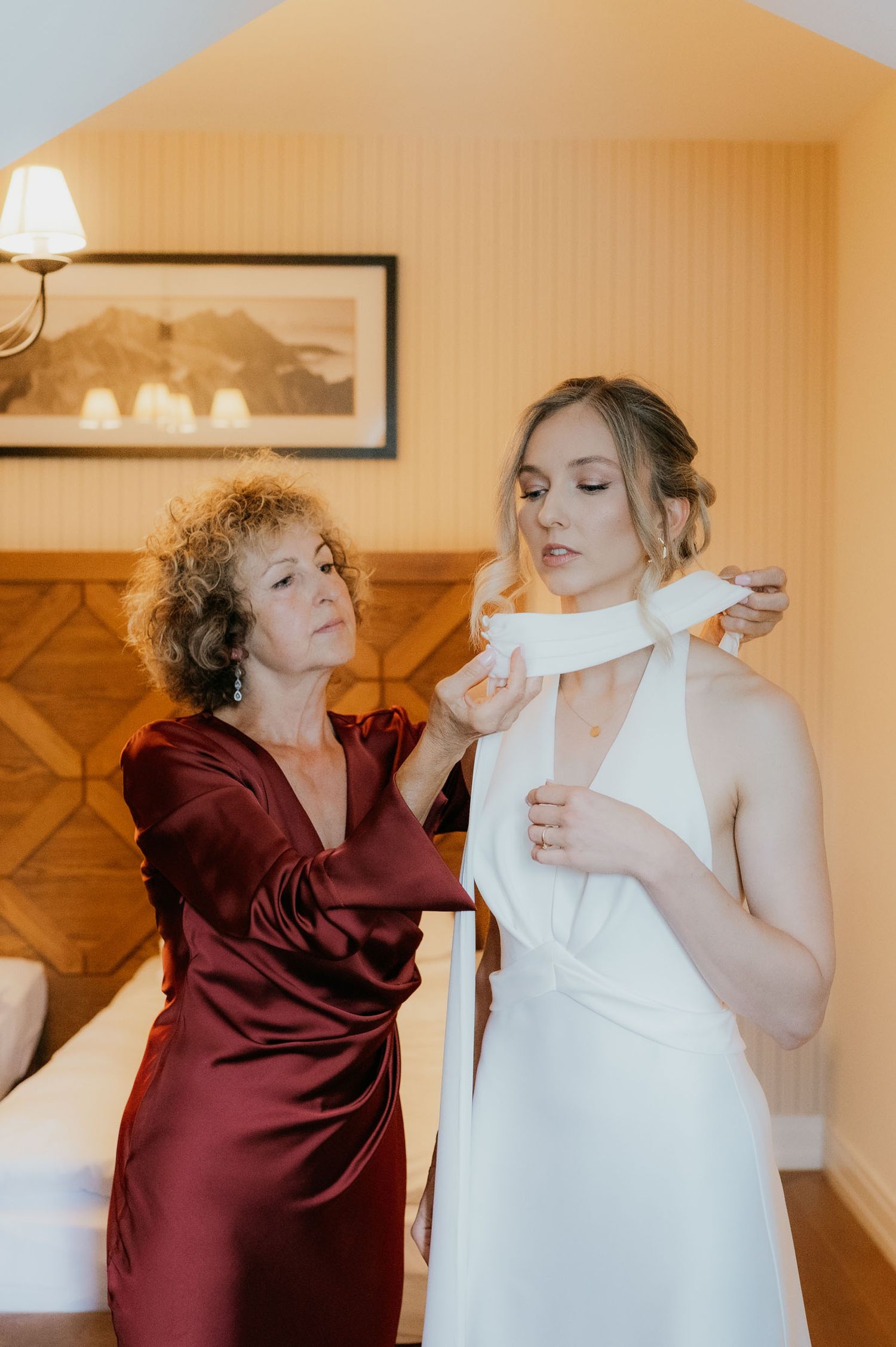 A bride in a white halter gown gets help tying her dress from a woman in a burgundy satin dress in a hotel room.