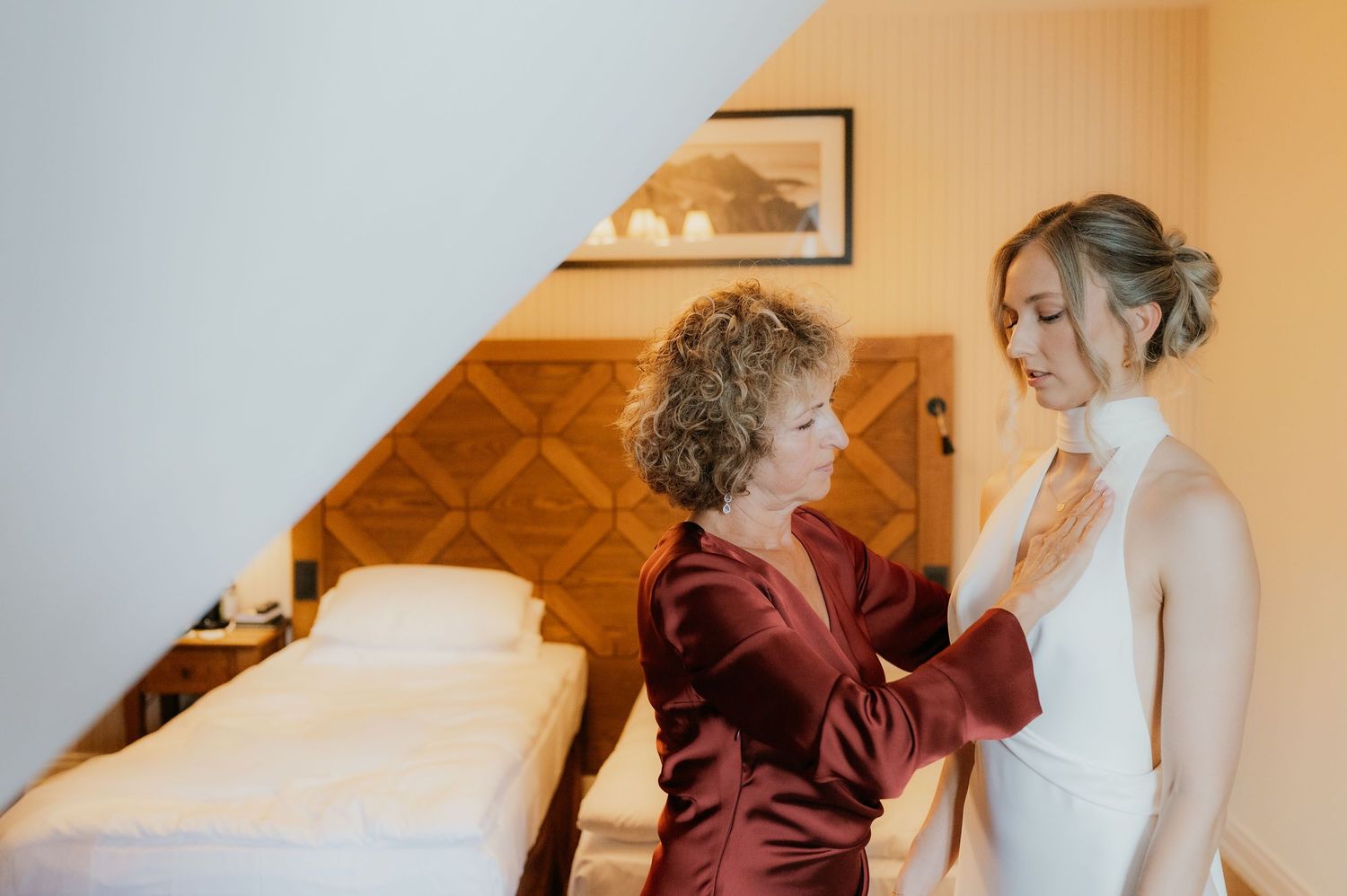 Mother helping bride in white gown prepare for wedding ceremony in cozy hotel room.