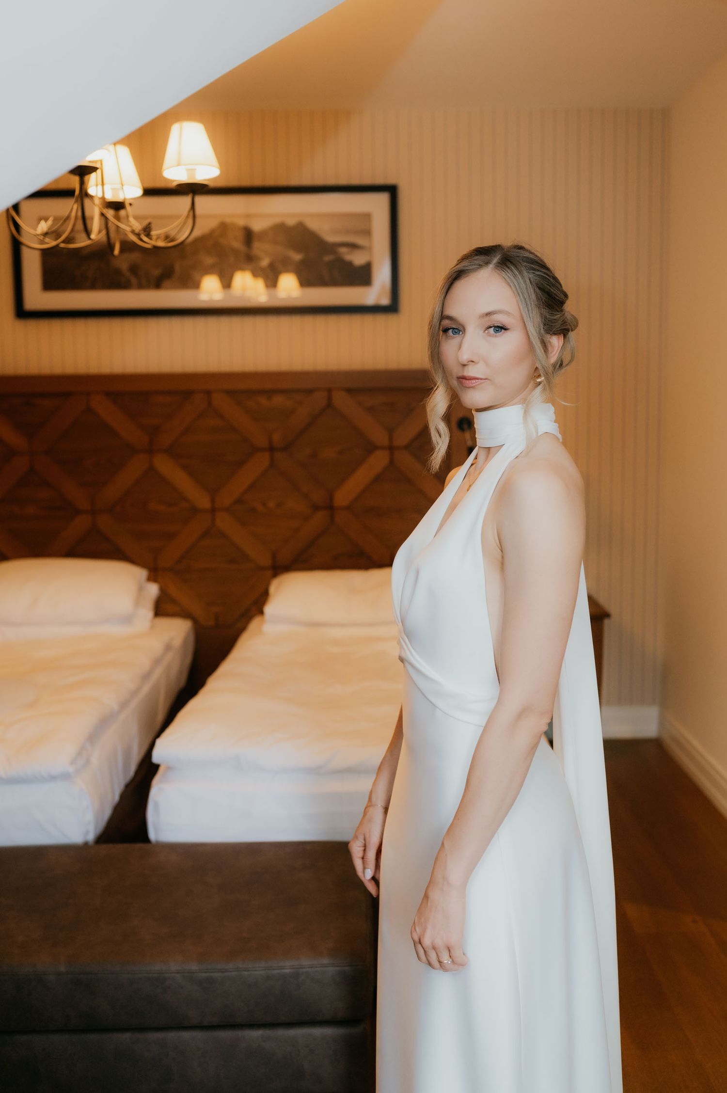 Bride in elegant white halter gown poses in a cozy hotel room with wooden headboard and warm lighting.