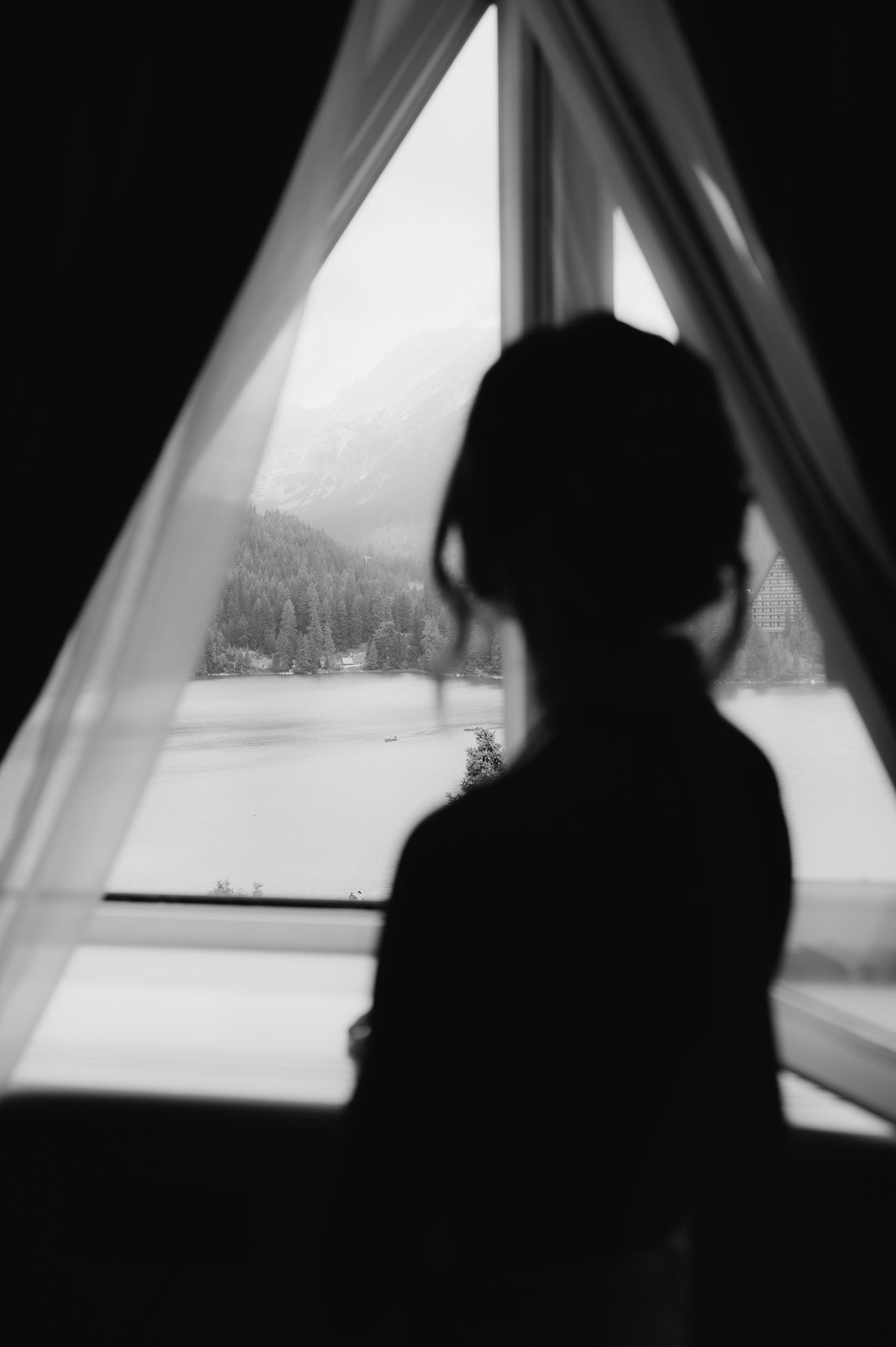 Silhouette of a woman gazing out a triangular window at a snowy mountain lake landscape in black and white.