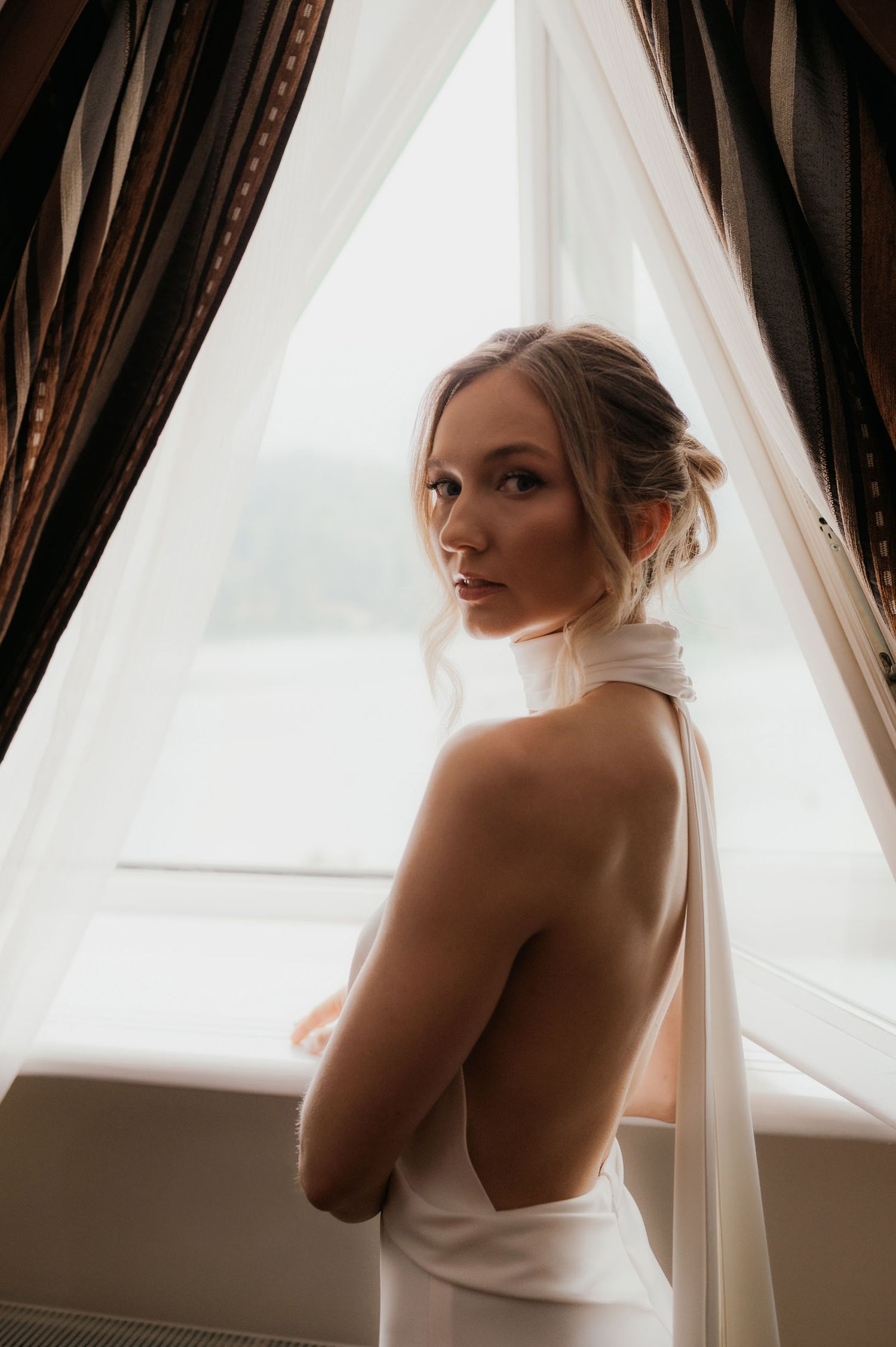 Elegant bride in a white backless halter gown poses by a draped window in soft natural light.