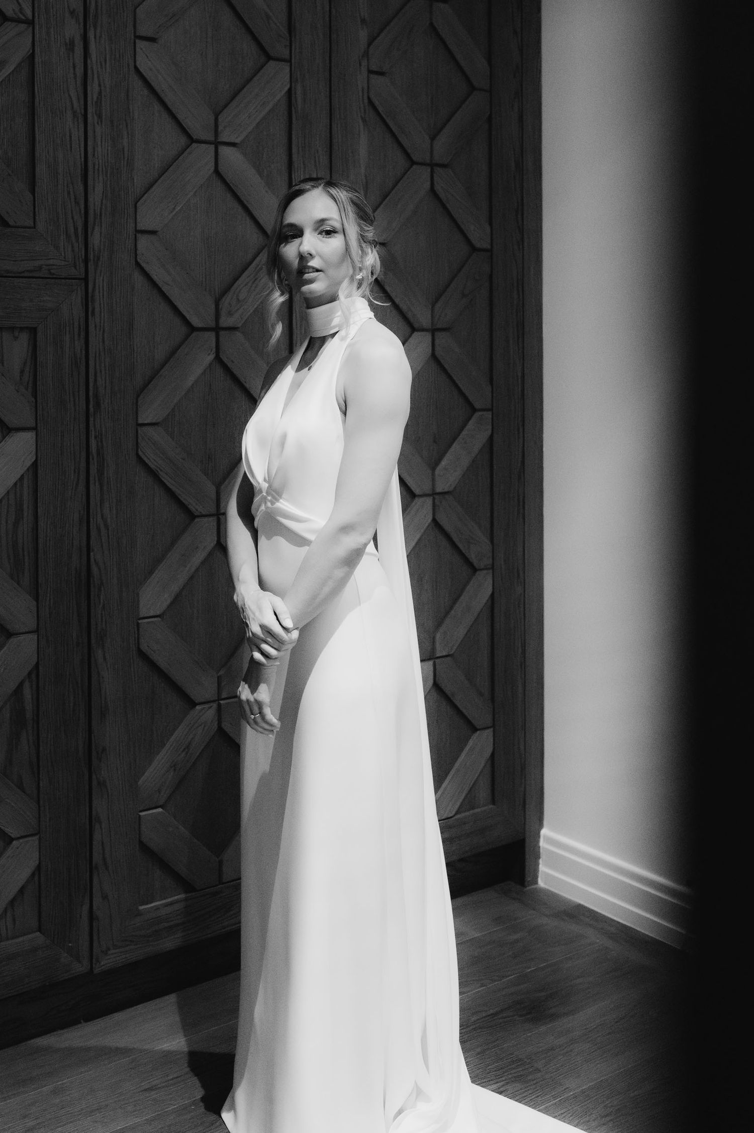 Elegant woman in white halter gown poses before geometric wood panel wall in black and white photo.