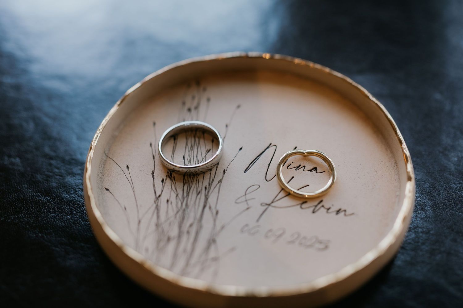 Two gold and silver wedding rings resting on a personalized ceramic dish engraved with names Nina and Robin.