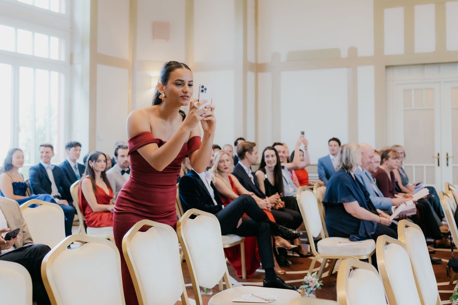 Woman in red dress photographs a wedding ceremony while guests in formal attire sit in white chairs.