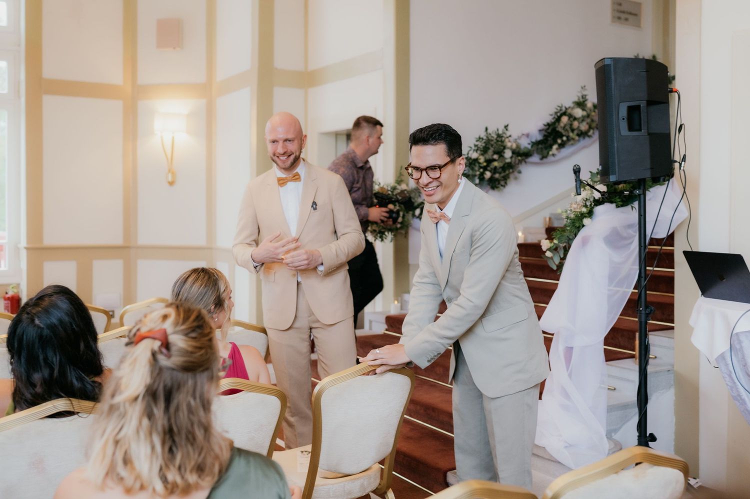 Two grooms in suits greeting wedding guests at an elegant indoor ceremony venue.