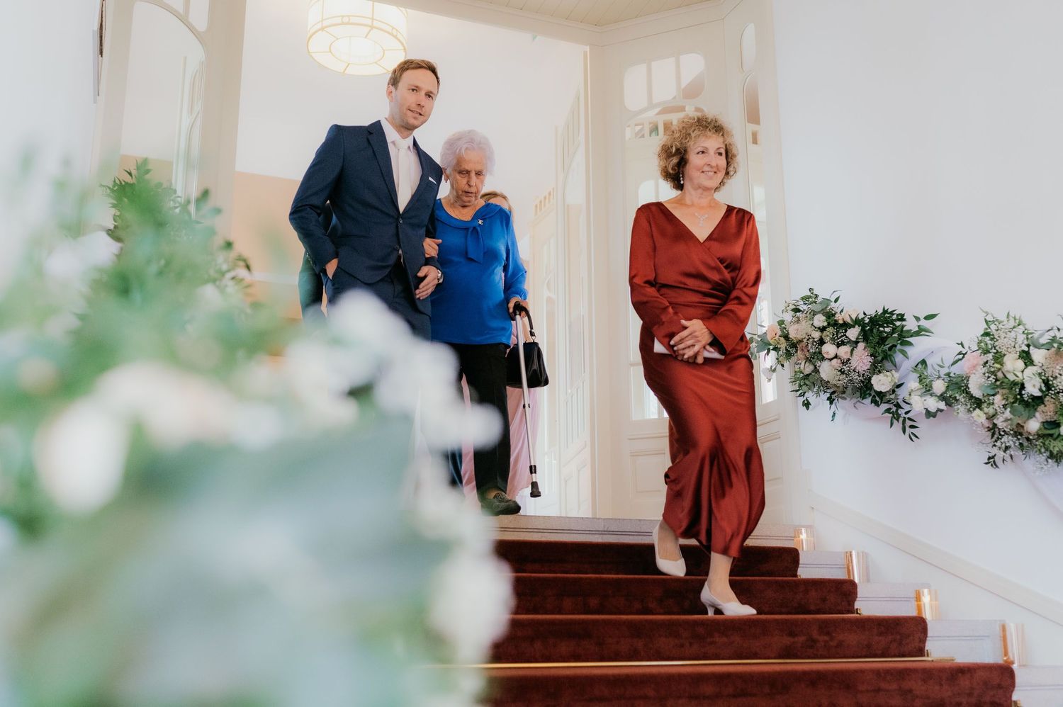Three elegantly dressed guests descend a grand staircase adorned with floral arrangements at a wedding venue.