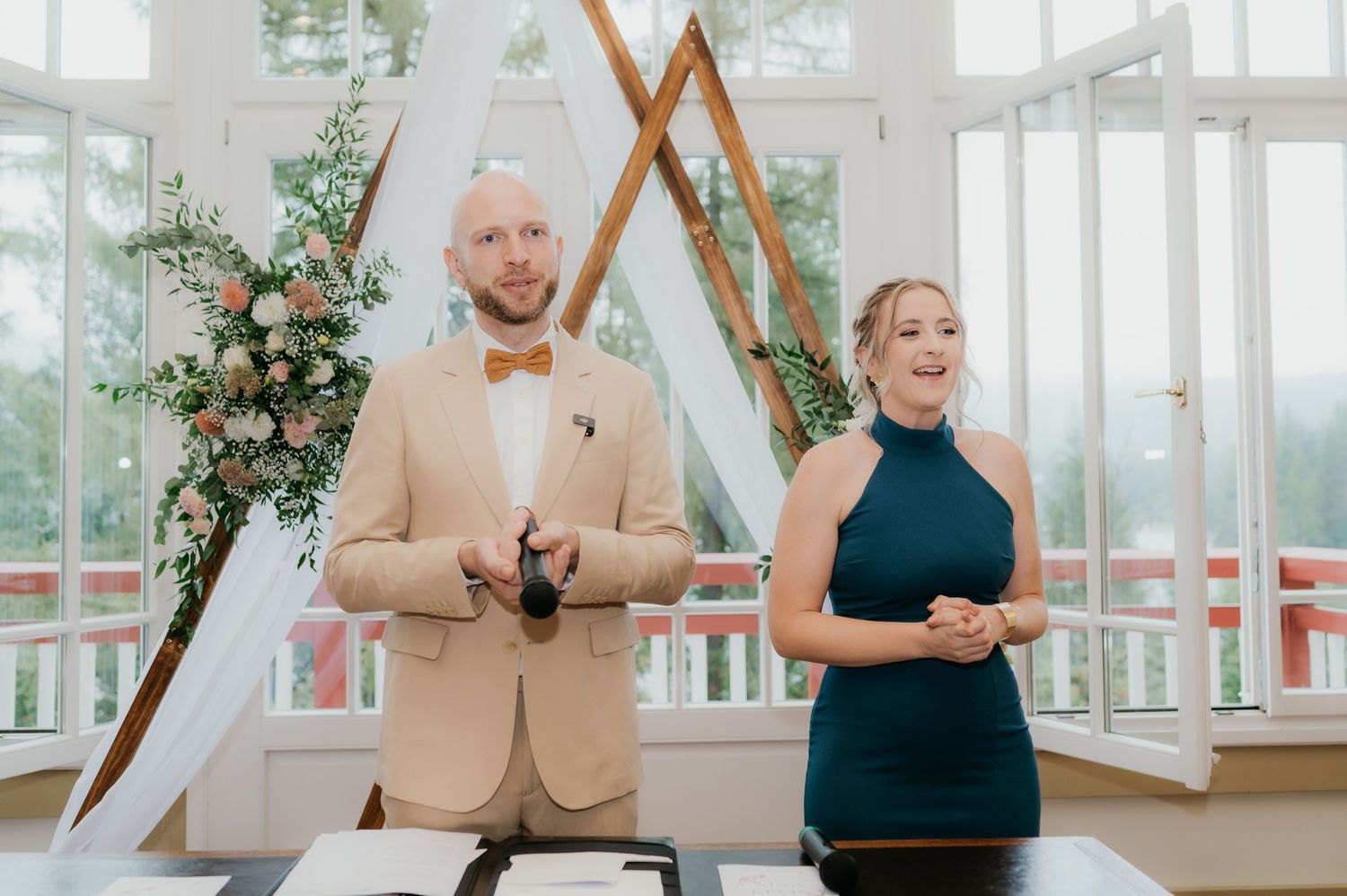 Man in beige suit and woman in teal dress stand at wedding ceremony altar with floral arch backdrop.