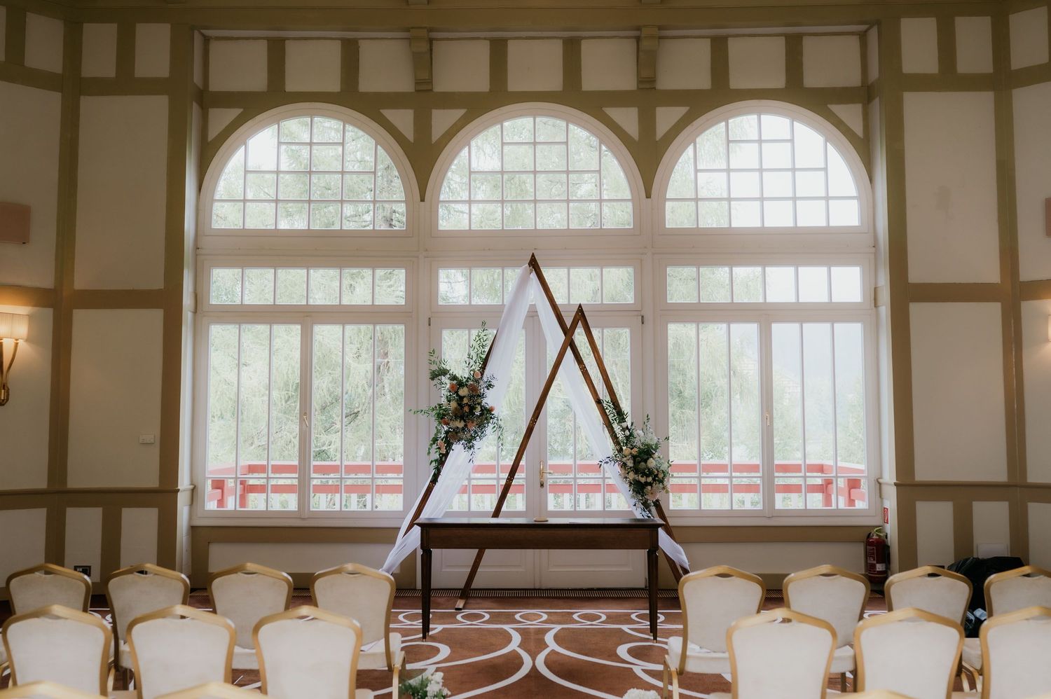 Elegant wedding ceremony setup with triangular wooden arch, floral accents, and white chairs in a bright ballroom.